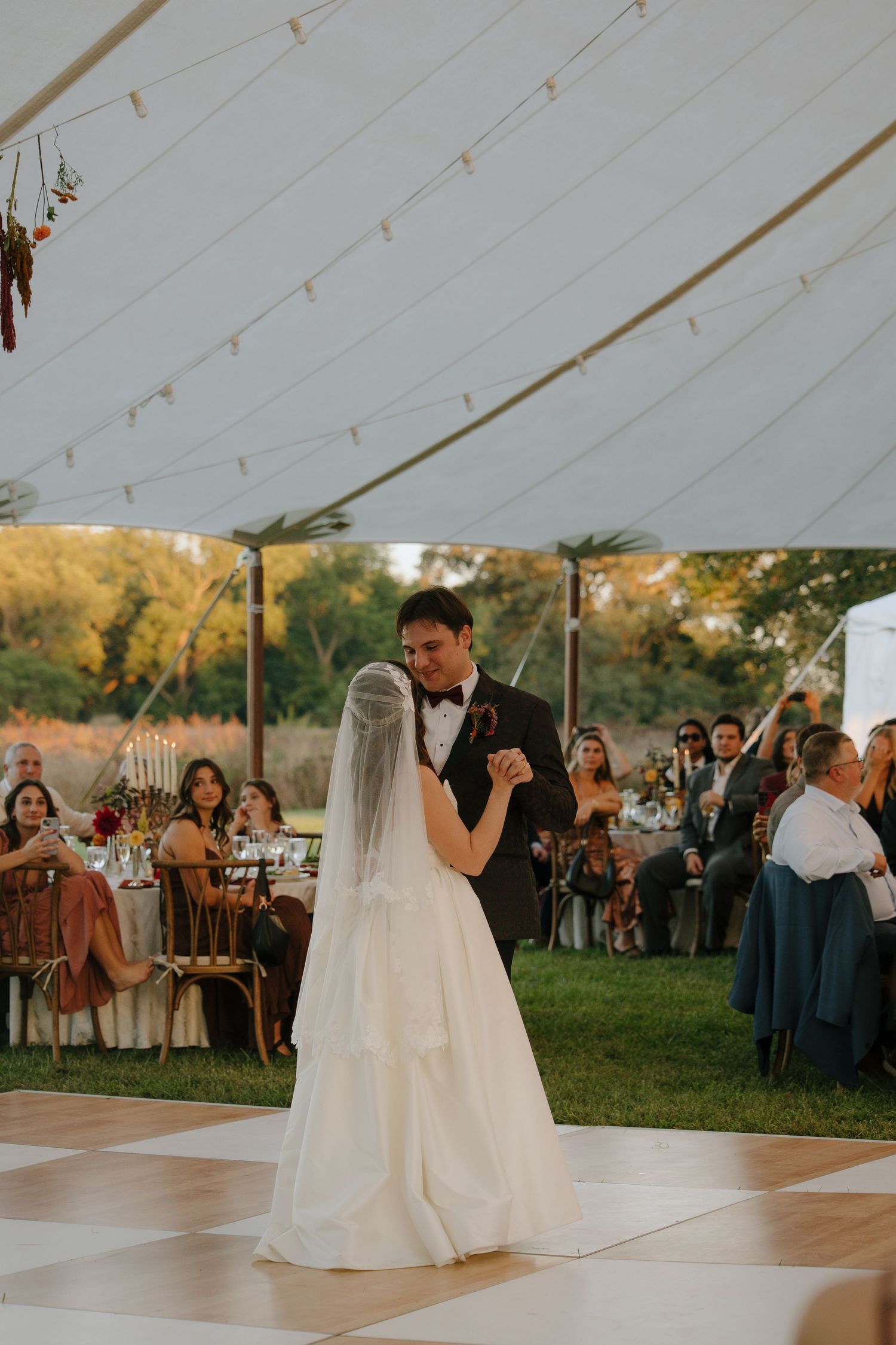Newlyweds share their first dance under an elegant white tent at sunset while guests watch from the sidelines.