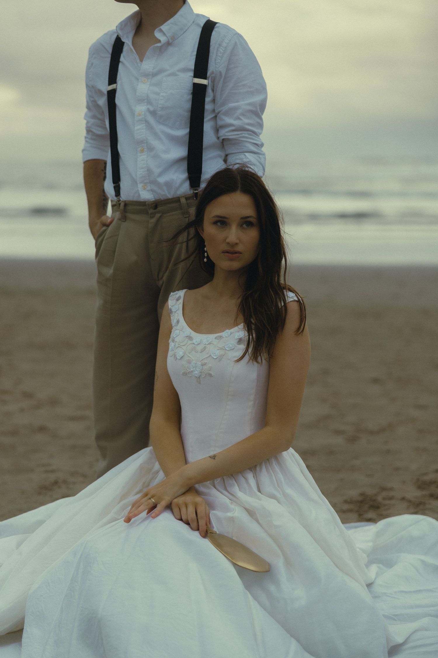 Bride in white gown sits on beach while groom in suspenders stands behind her, with ocean waves in the background.