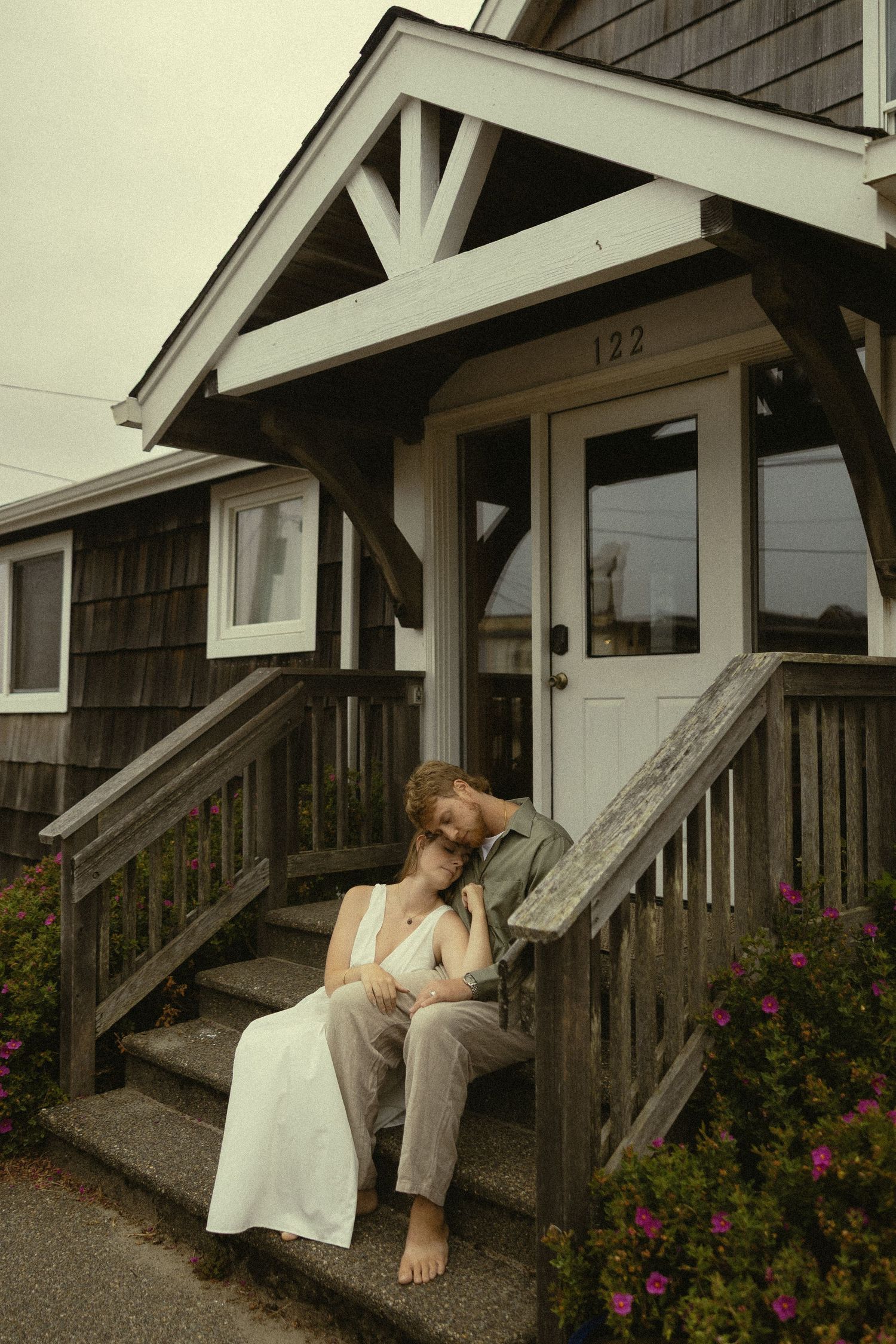 Couple sitting on wooden porch steps of cottage 122, sharing a tender embrace in casual elegant attire.