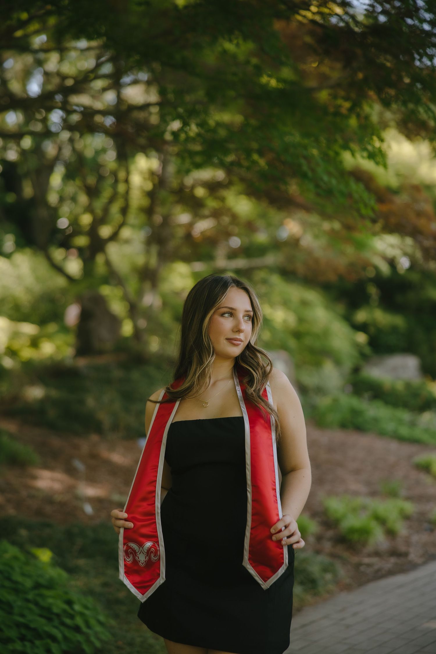 Young woman in black dress wearing red graduation stole poses in lush green garden for senior portrait.