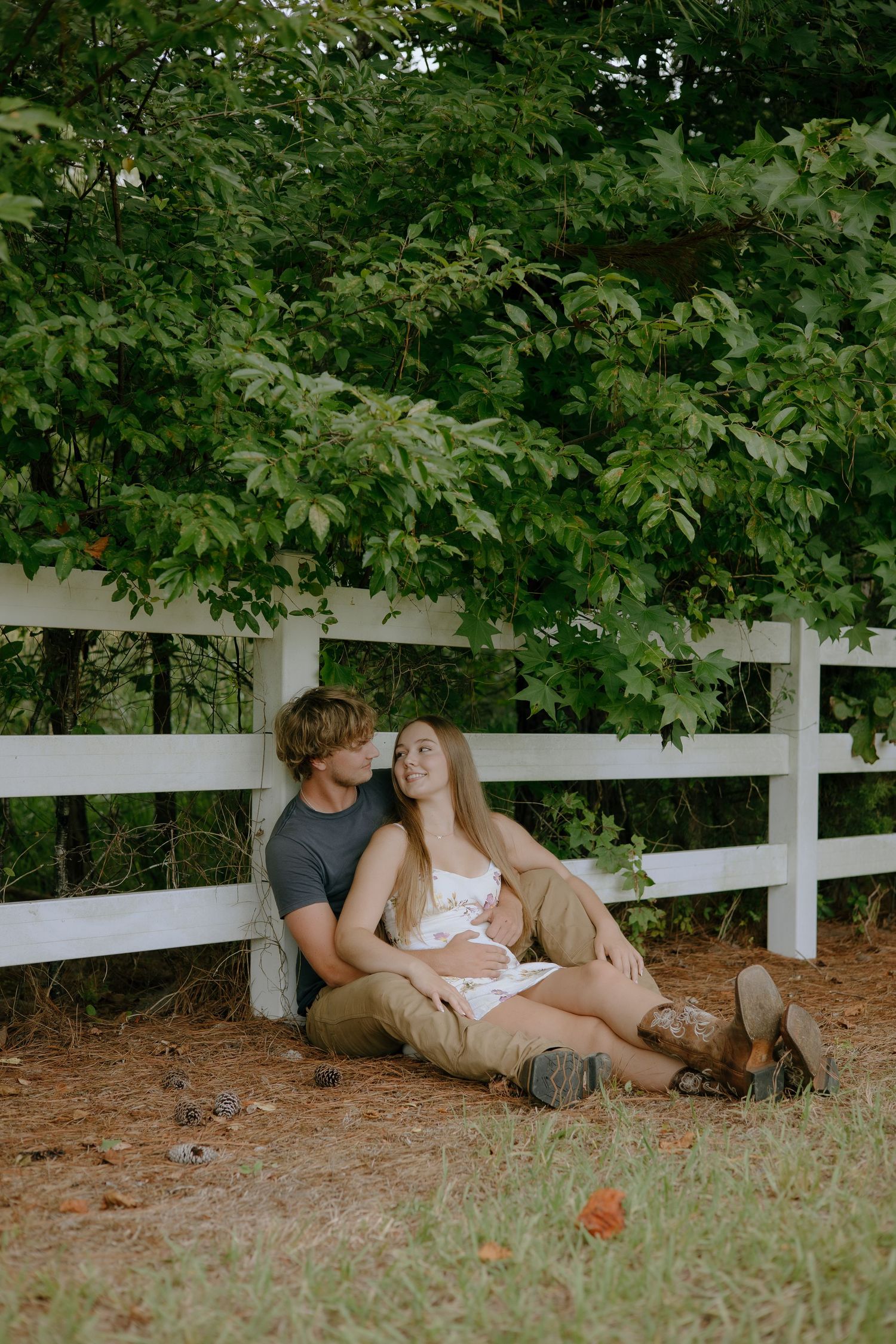 A couple sits together against a white fence surrounded by lush green trees, sharing a romantic moment outdoors.