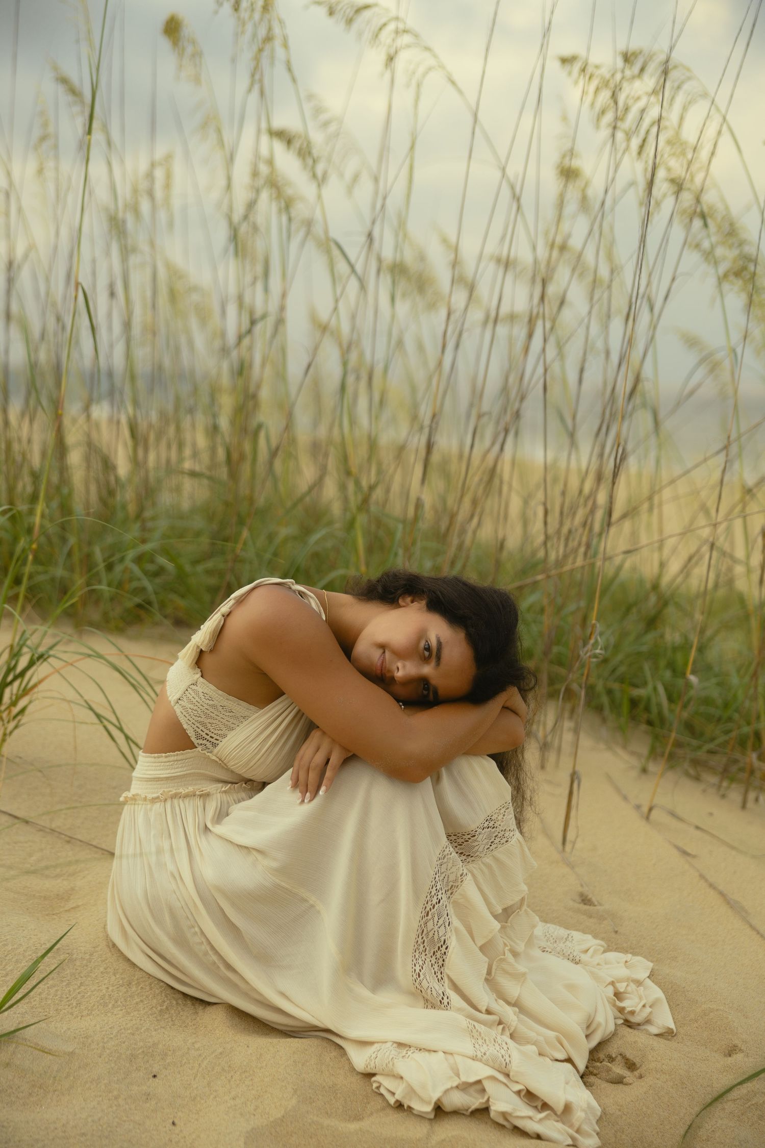 Woman in white dress sitting on sand dunes among sea oats at beach.