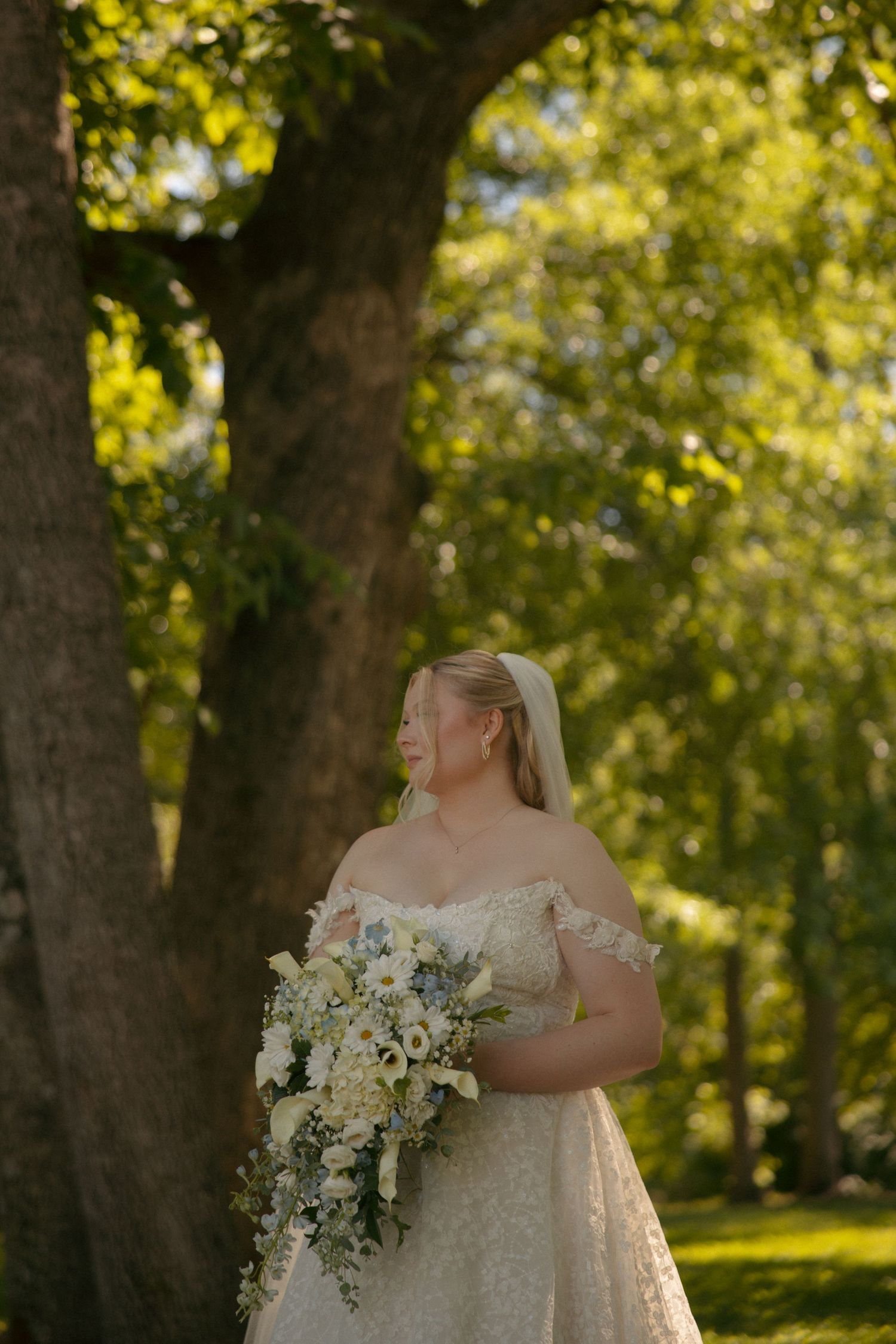 Bride in off-shoulder lace gown holds cascading white bouquet outdoors under lush green trees on her wedding day.