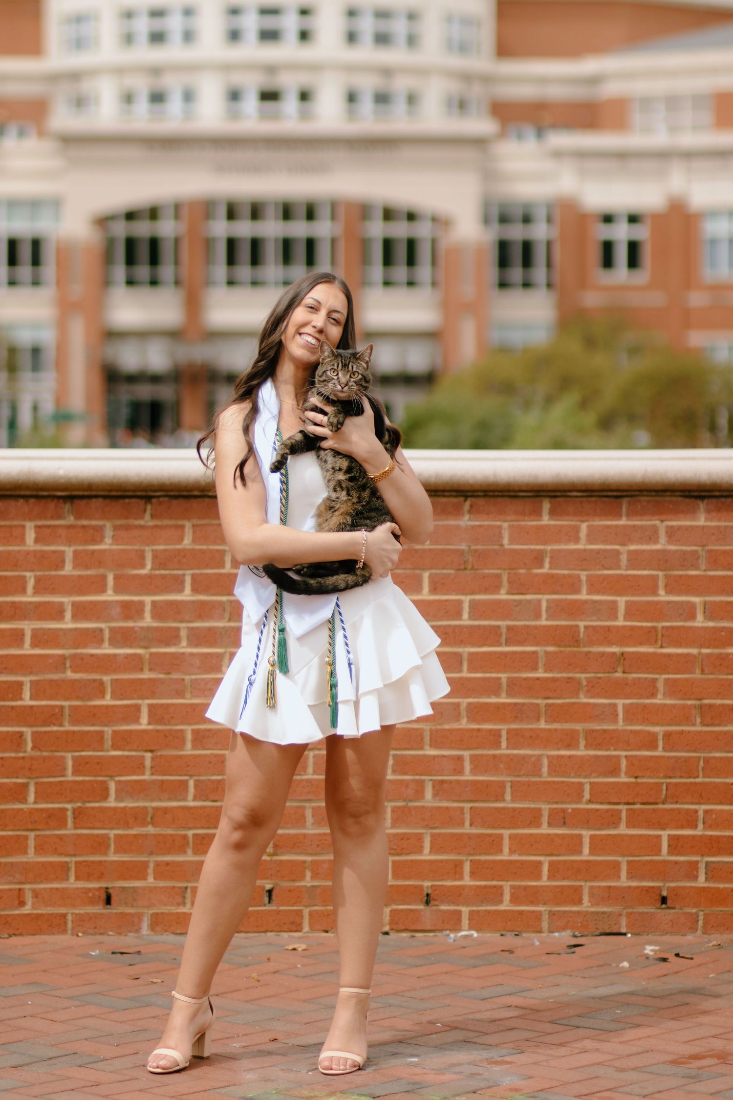 A person in a white dress holds and plays with a tabby cat near a brick wall.