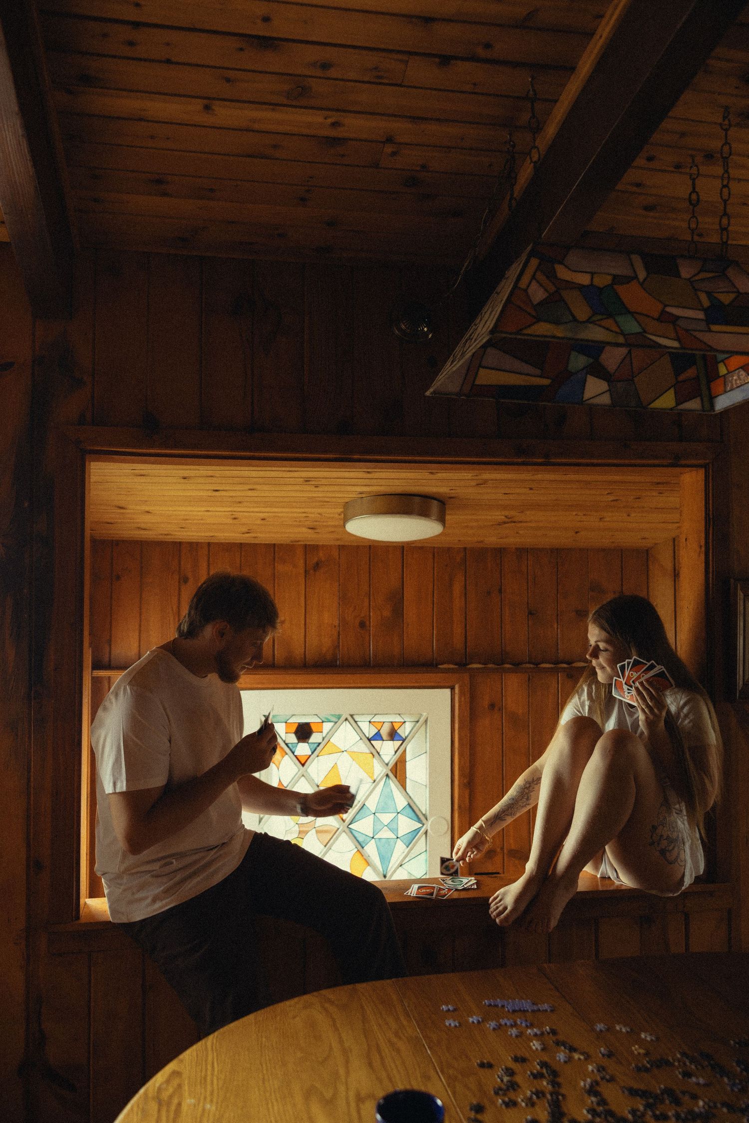 Couple doing a jigsaw puzzle together in a cozy wooden cabin with warm lighting and stained glass window.