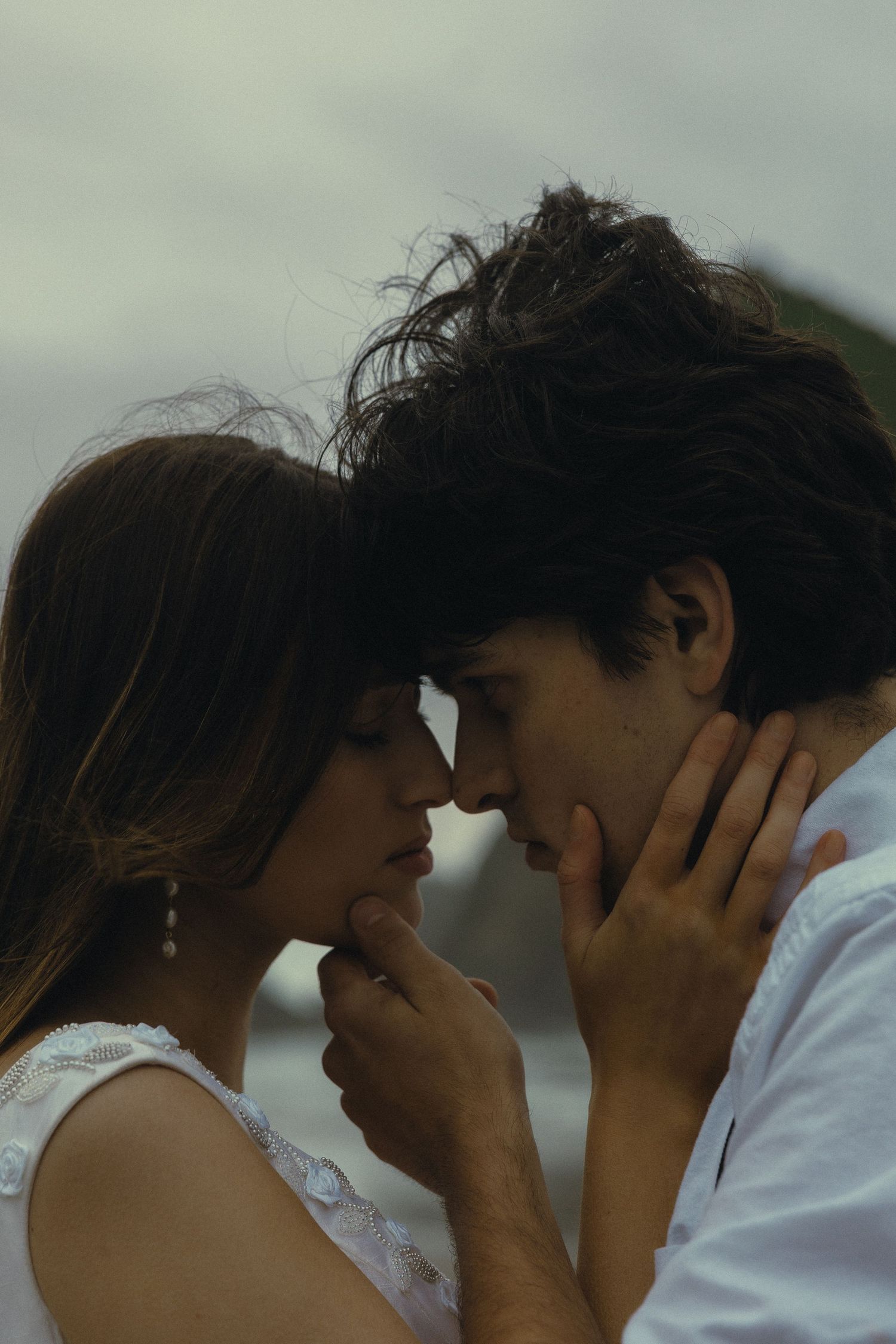 A couple shares an intimate moment, foreheads touching, hands gently cradling each other's faces against a cloudy sky.