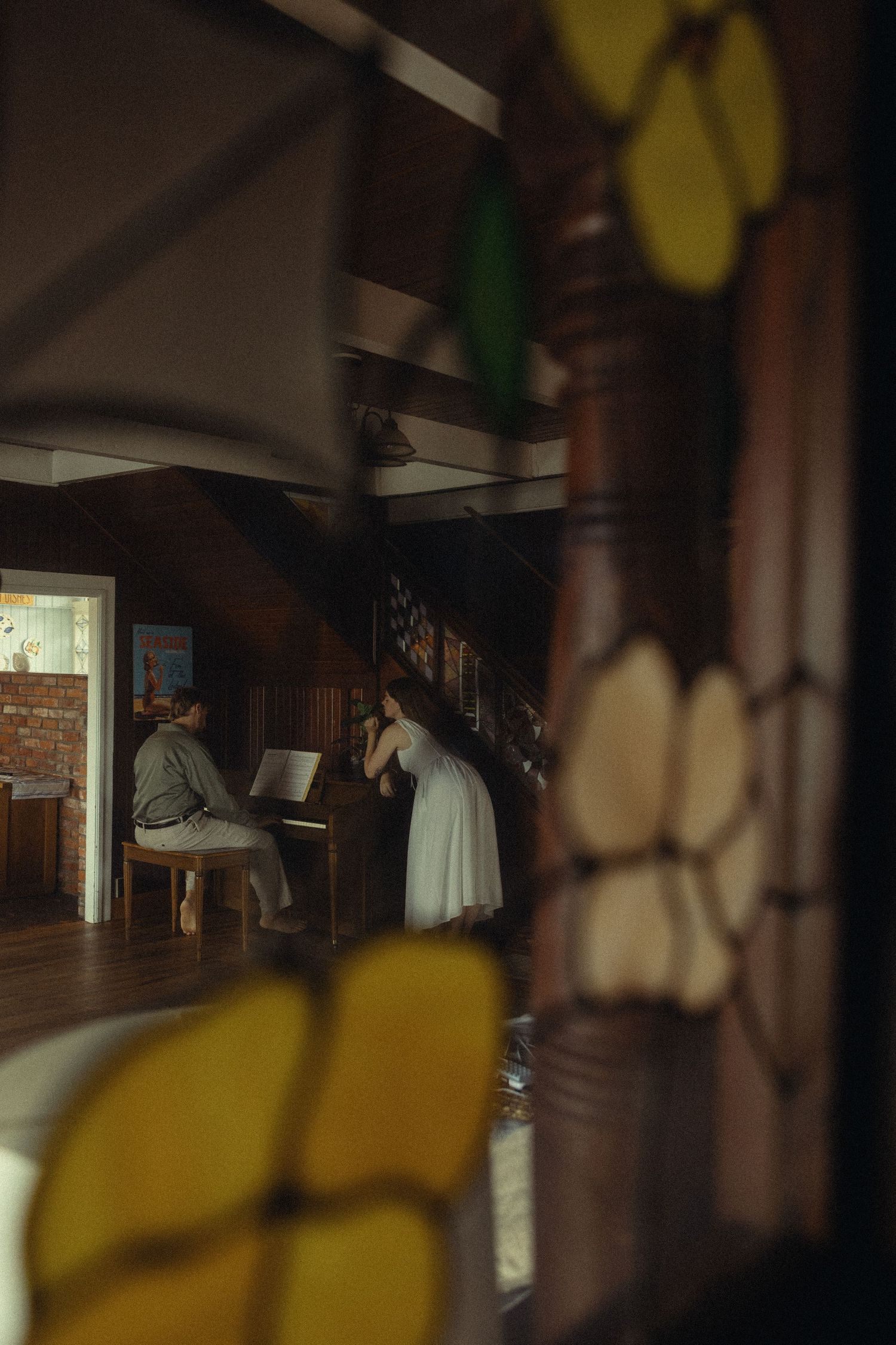 A couple in a rustic wooden interior, one playing piano while the other stands nearby, viewed through stained glass.