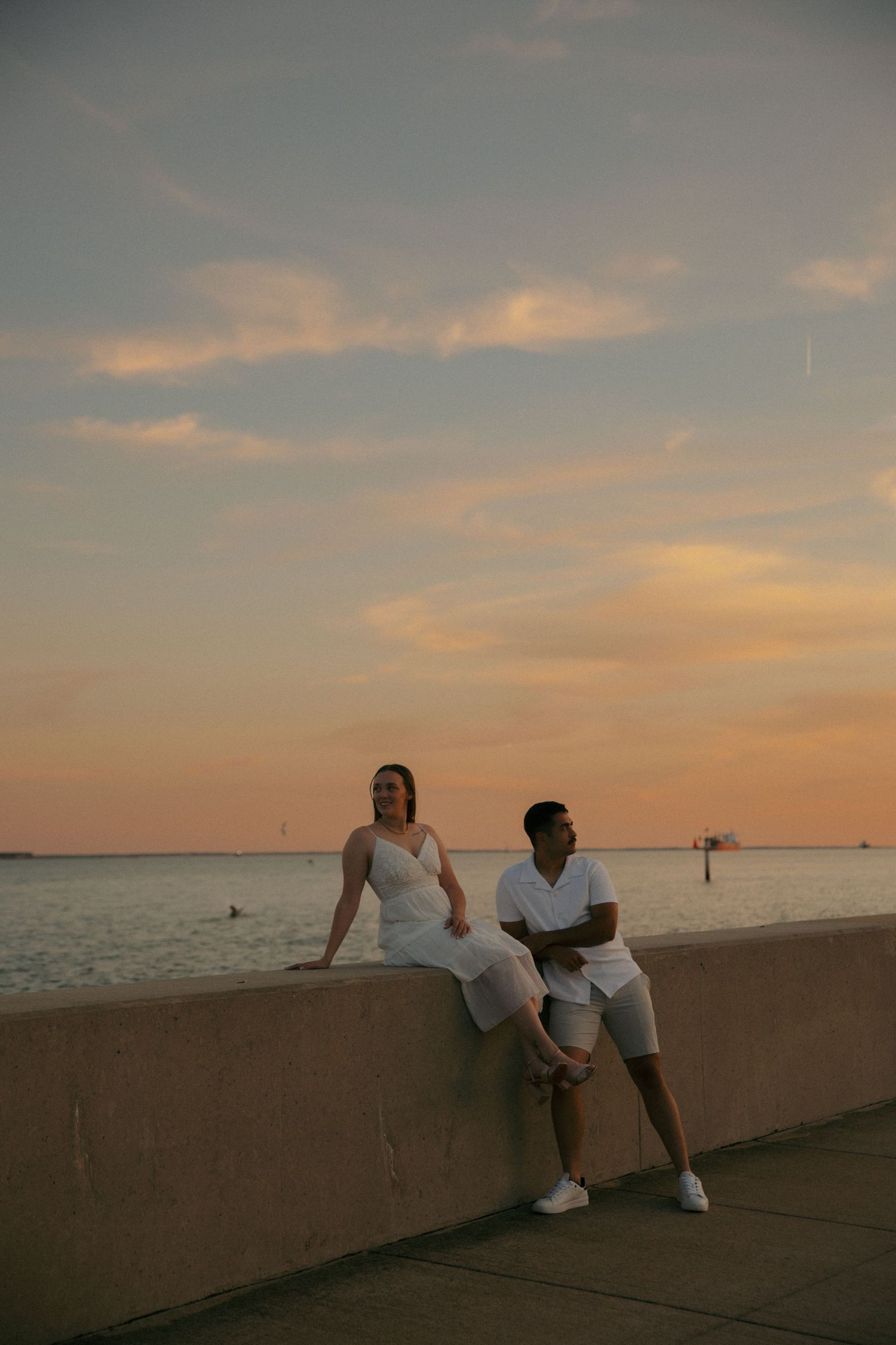 Couple in white outfits sitting on a waterfront wall at sunset with a golden sky and calm sea behind them.