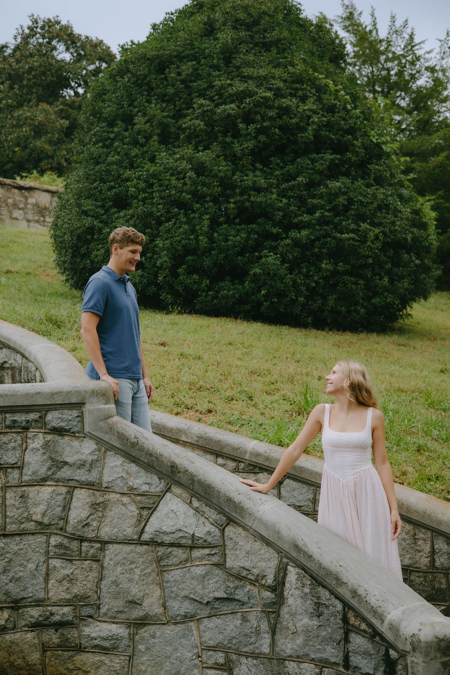 A couple shares a romantic moment on stone steps surrounded by lush green trees and grass.