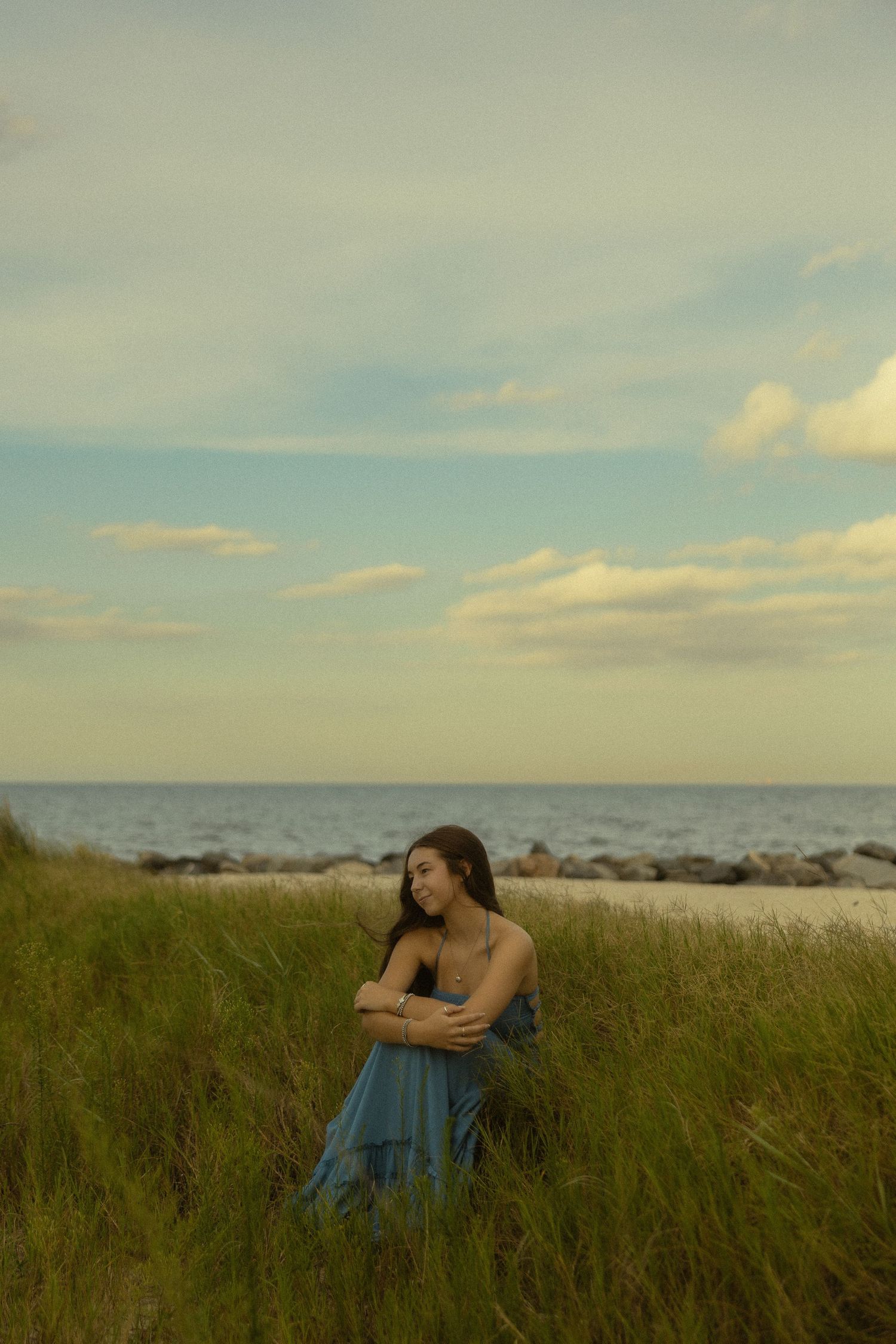 Young woman in blue dress standing in tall grass by the ocean shore on a cloudy day.