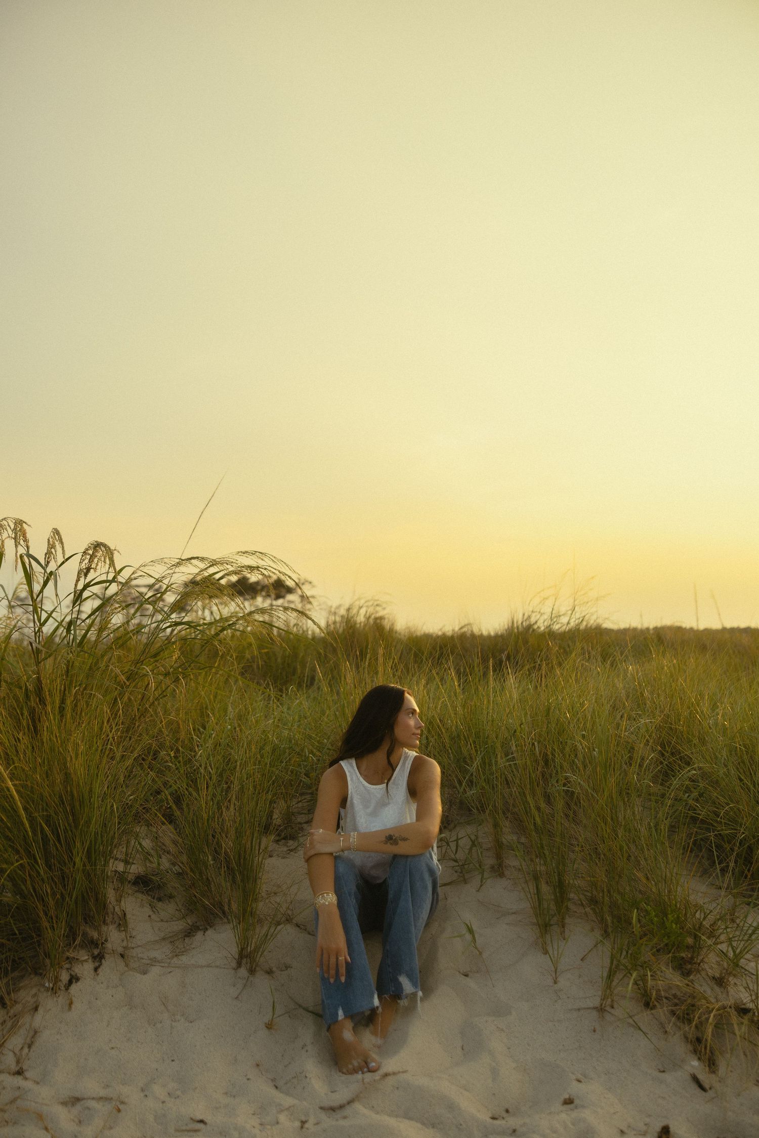Woman sitting on sandy beach dunes at sunset, surrounded by tall grass, wearing white tank top and jeans.