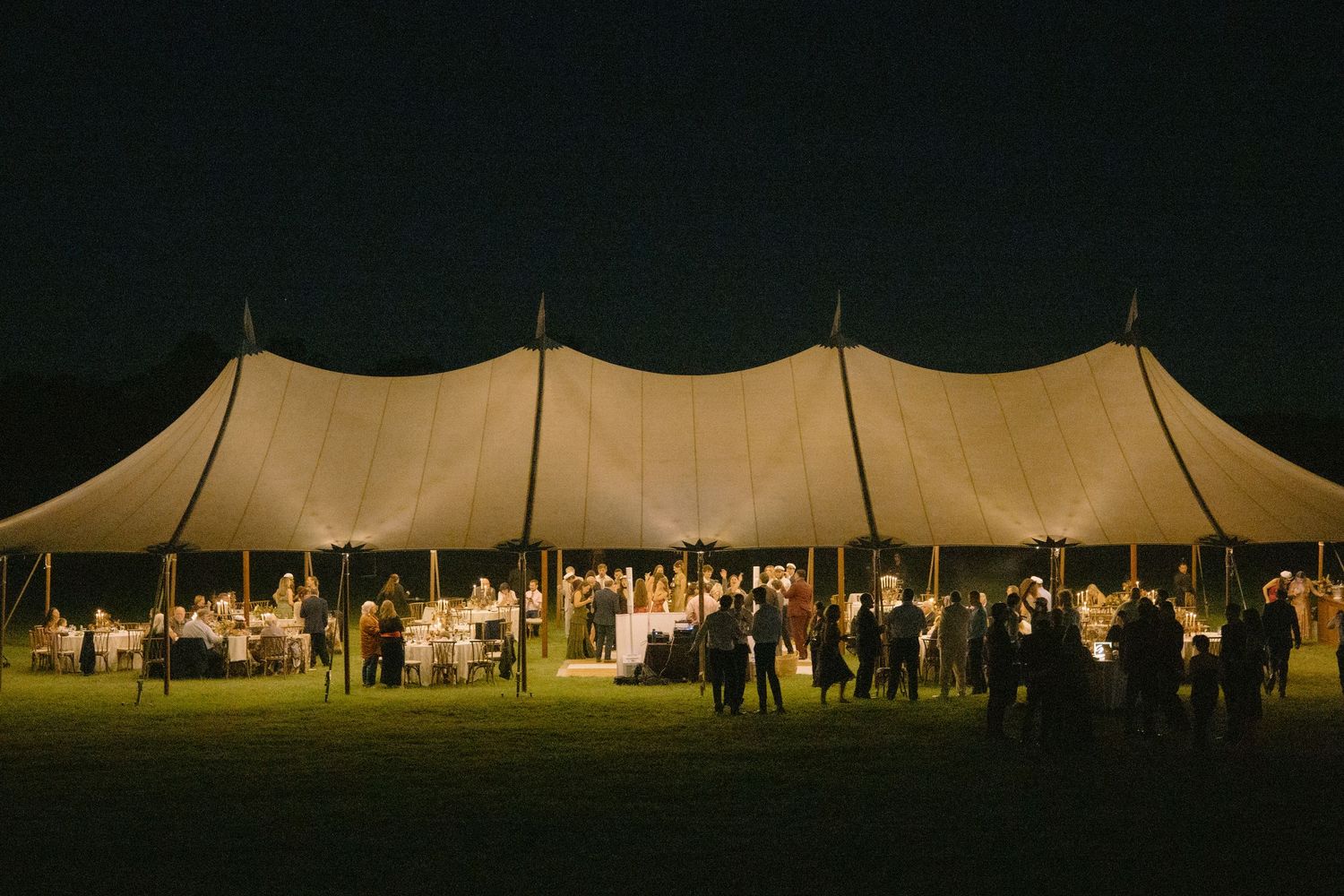 A large beige wedding tent glows warmly at night with guests gathered underneath for an outdoor reception celebration. Piney Grove, Maryland