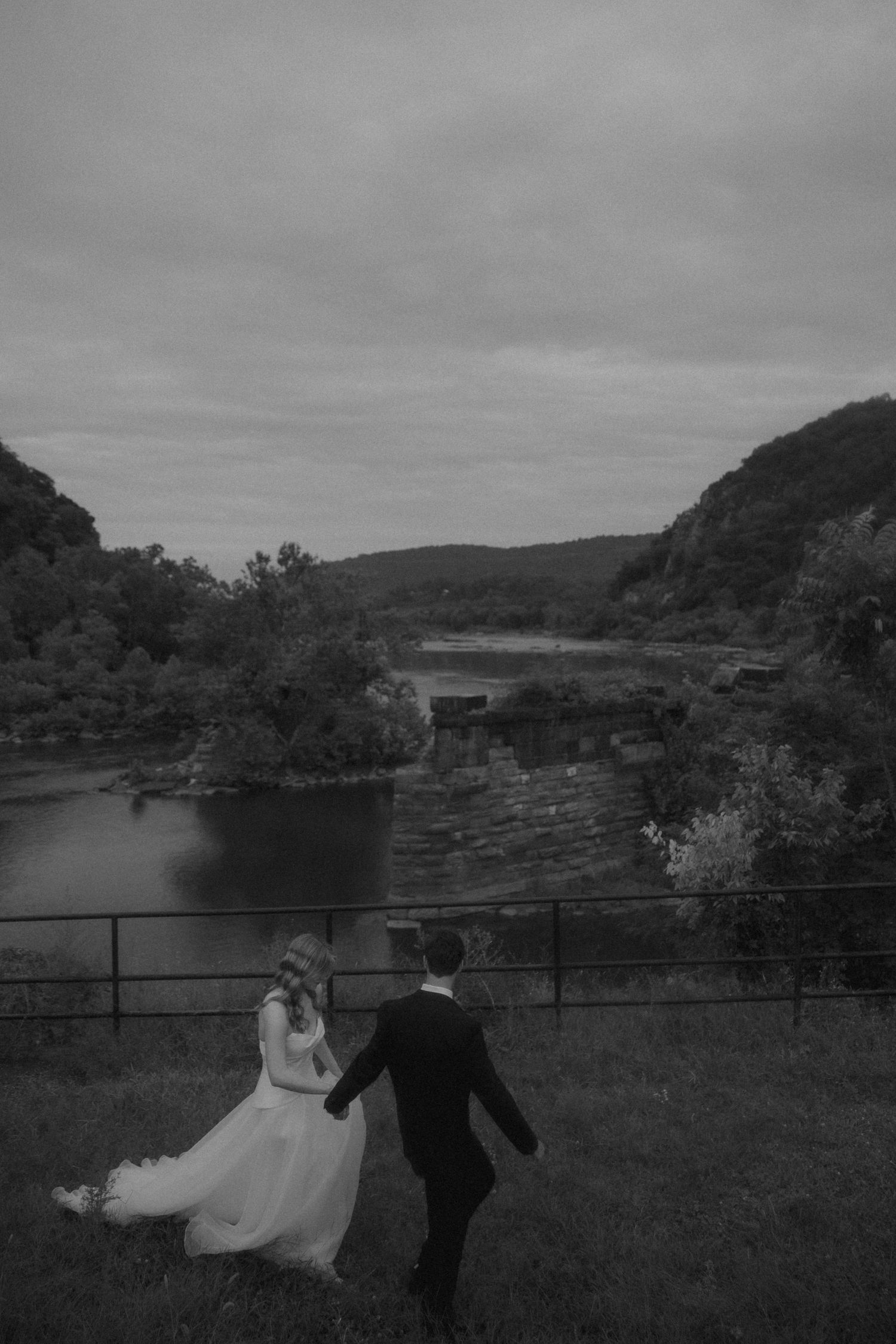 Black and white photo of bride and groom holding hands overlooking a scenic river valley on their wedding day.