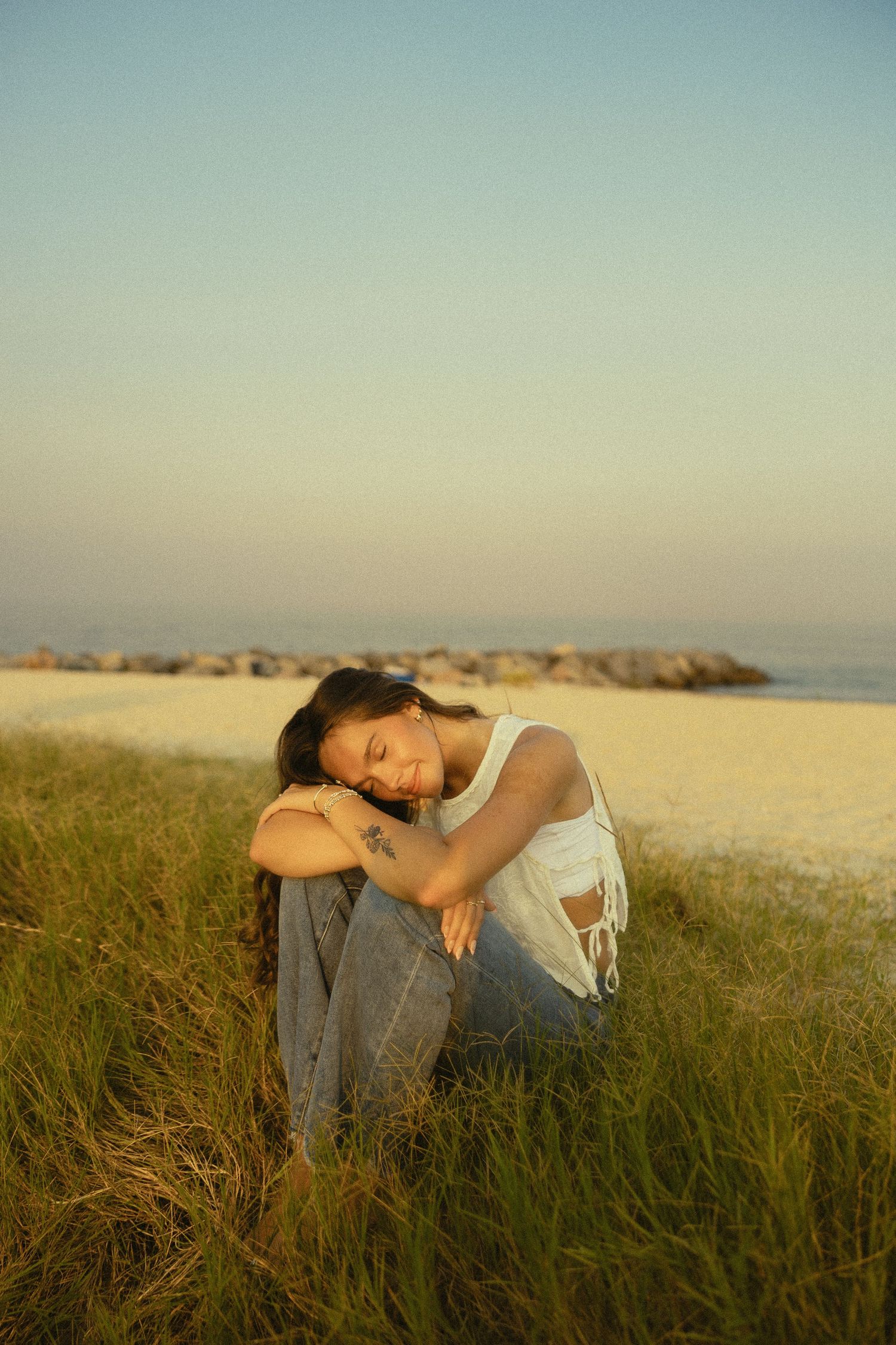 Young woman sitting in beach grass at sunset, hugging her knees in a peaceful, contemplative pose near the shoreline.