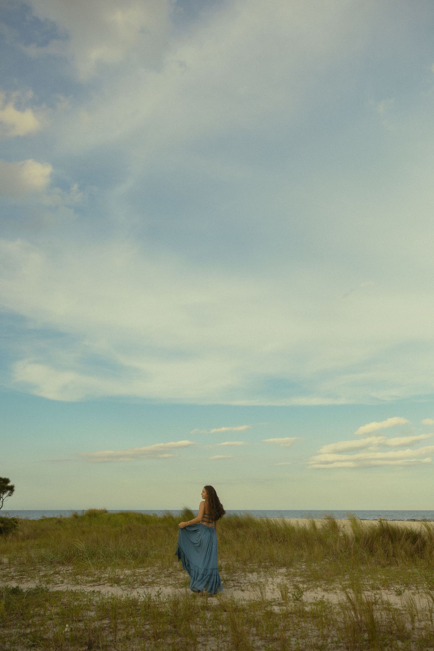 Woman in blue skirt stands on sandy beach dunes gazing at the ocean under a vast cloudy sky.