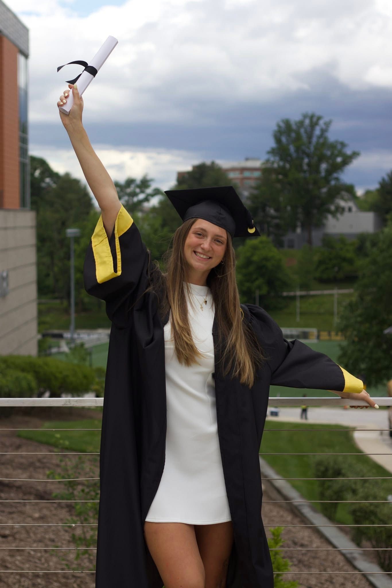 Smiling female graduate in black cap and gown holding diploma aloft outdoors on a cloudy day.