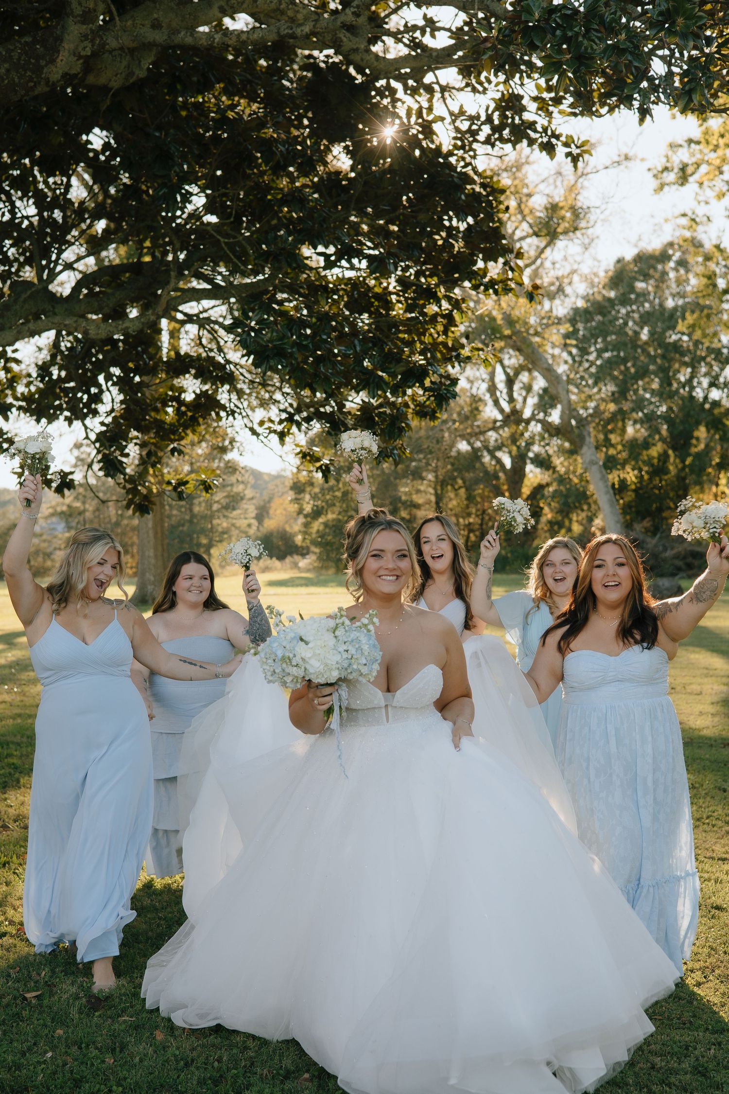 A group in white dresses and gowns pose together outdoors during golden hour under trees in a natural setting.