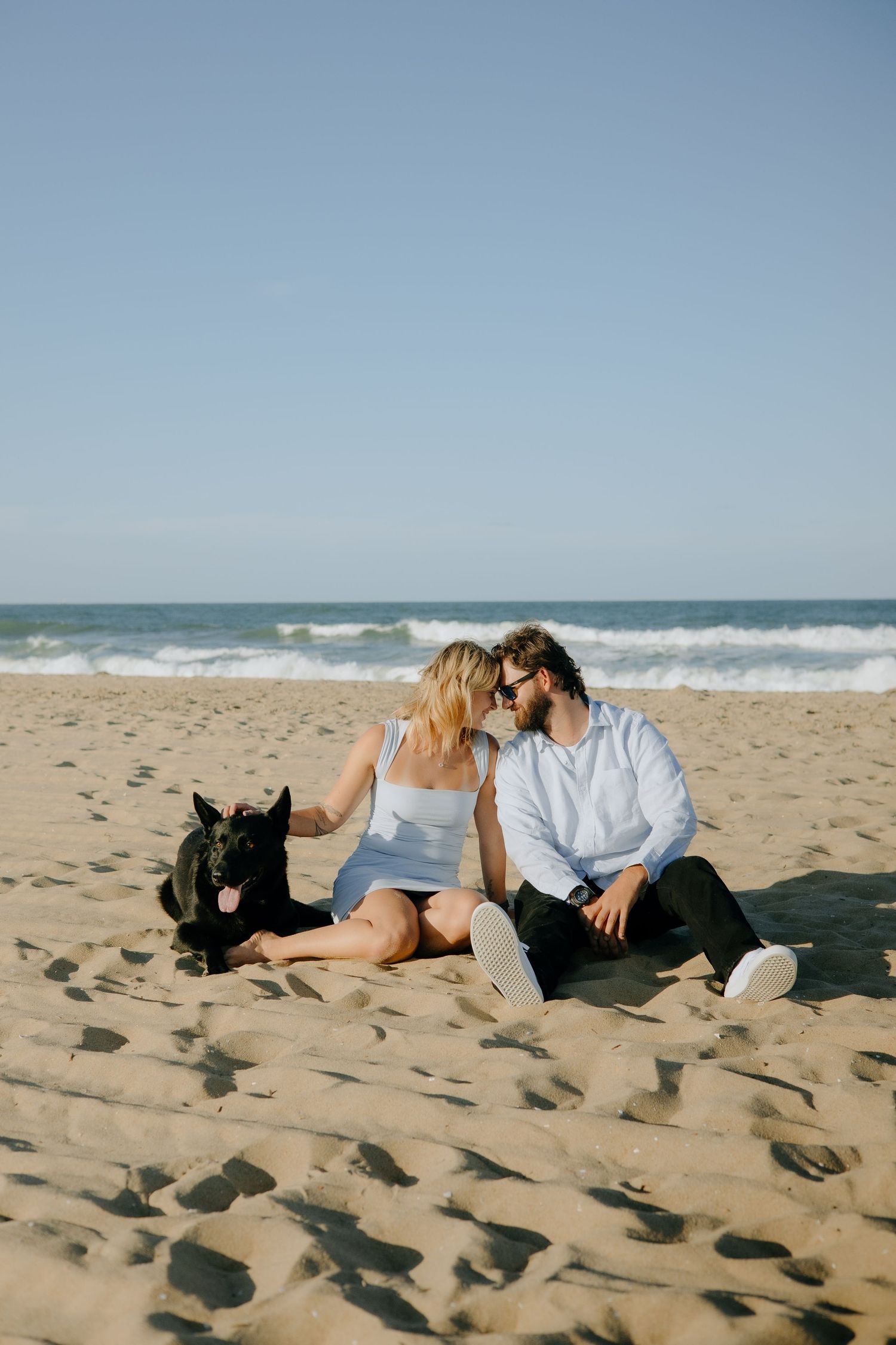 A couple lounges on the beach sand with their black dog beside them.