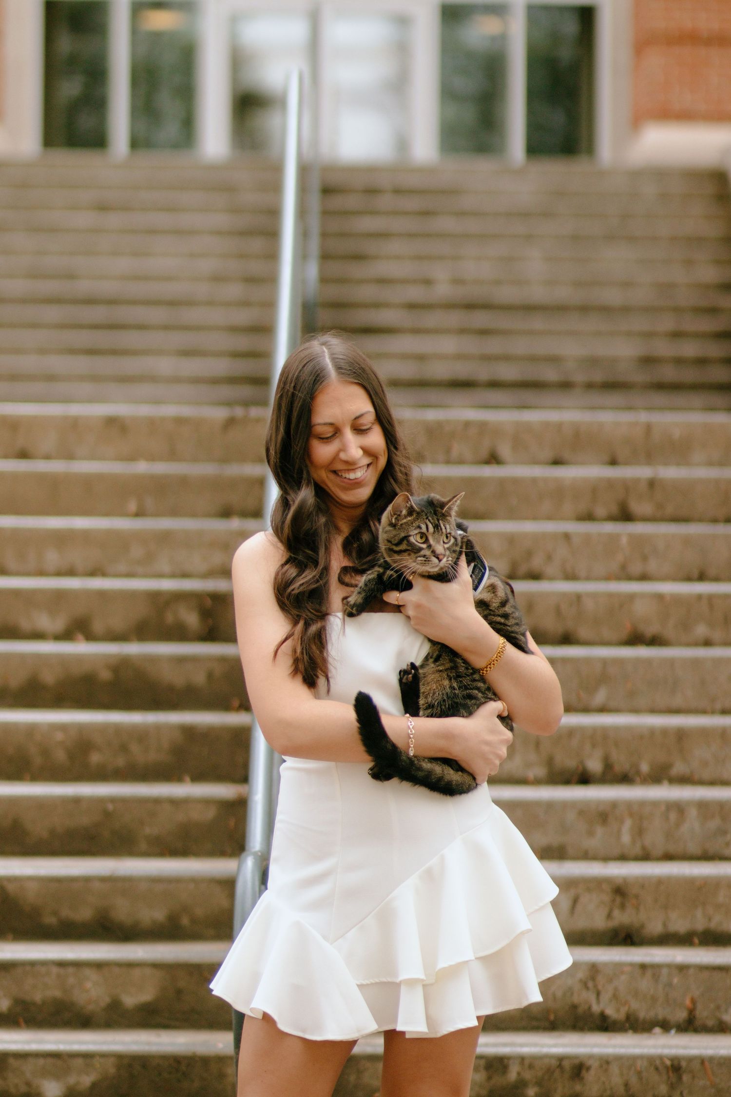 A person in a white dress sits on concrete steps while holding a tabby cat.