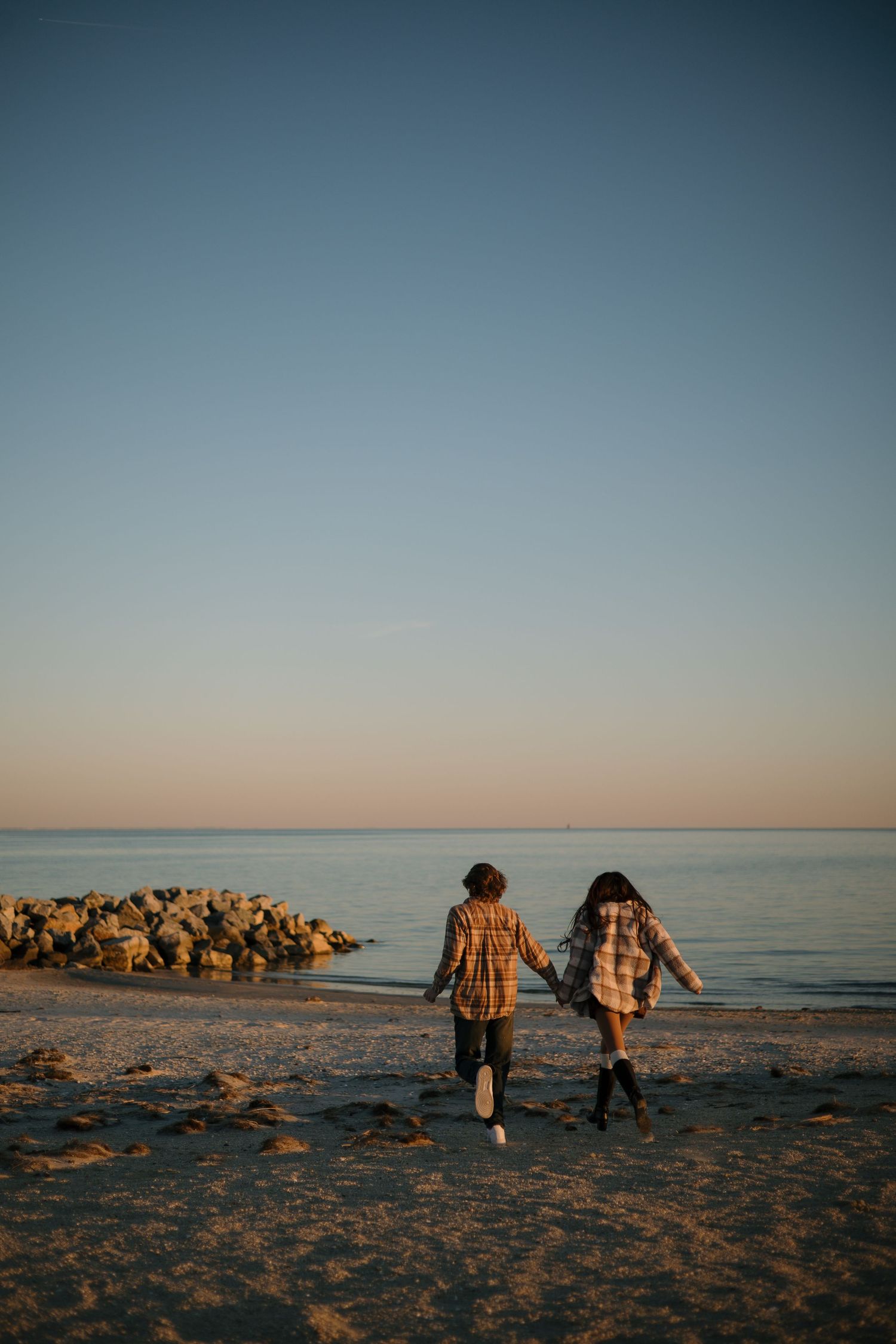 A couple walks along a scenic beach at sunset with large rocks visible in the distance.