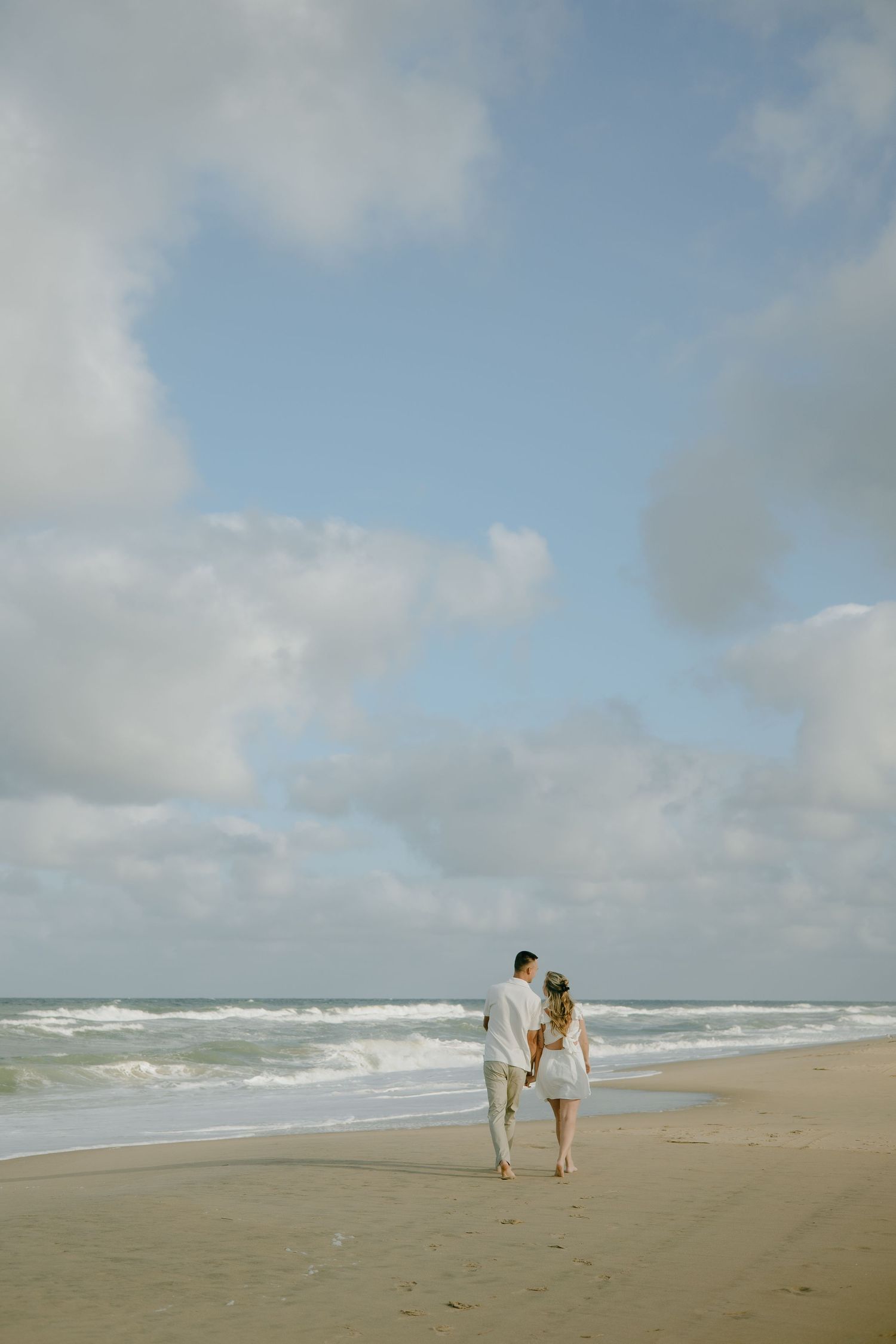 A couple walks together on a scenic beach with crashing waves and cloudy skies.