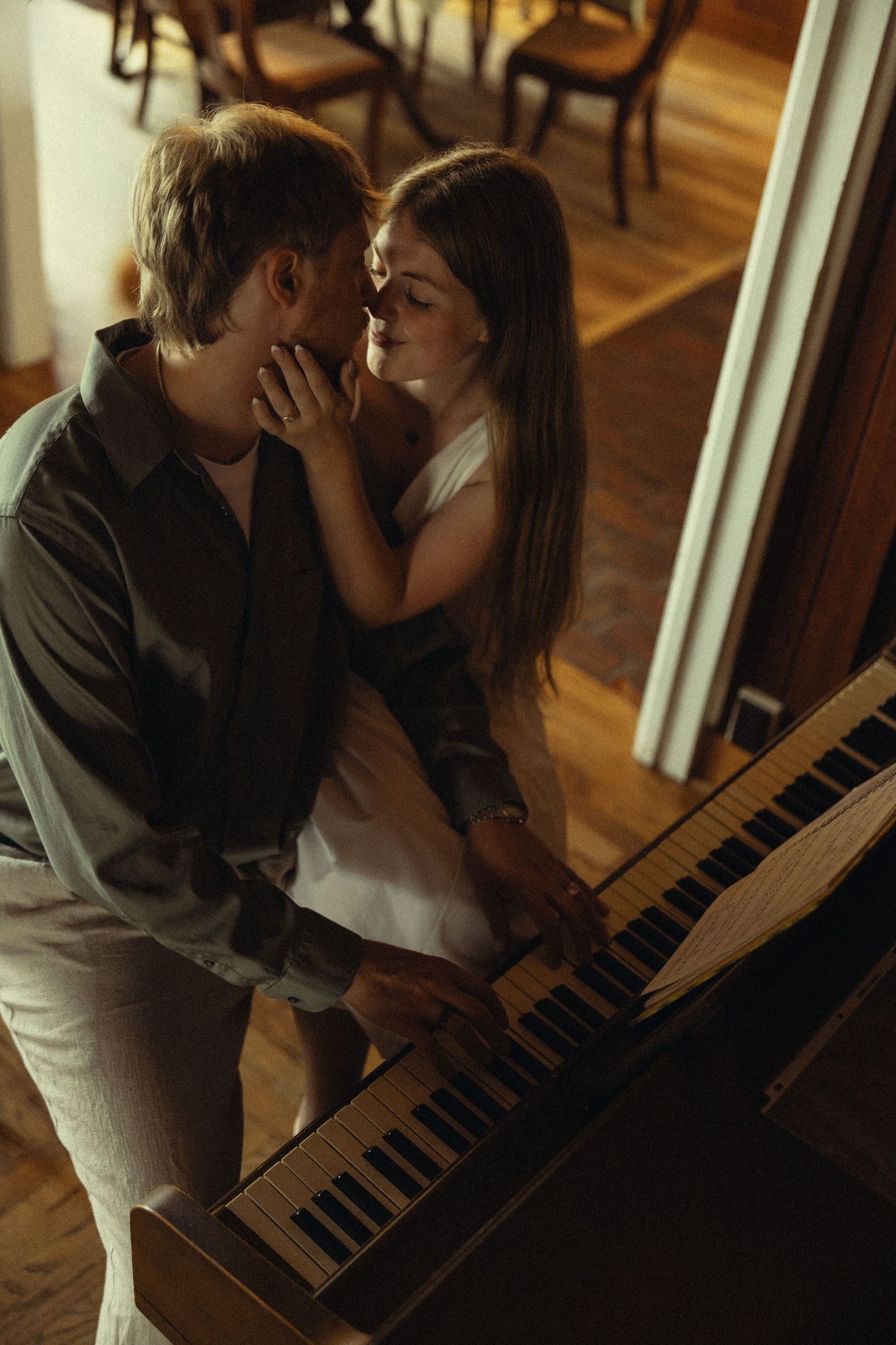 Romantic couple sharing an intimate moment at a piano in a warmly lit room with hardwood floors.