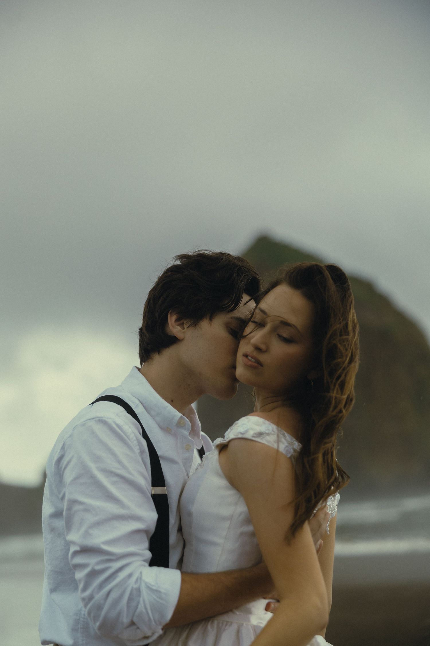 Couple sharing a romantic embrace on a moody beach, man in suspenders kissing woman in white dress near Haystack Rock.