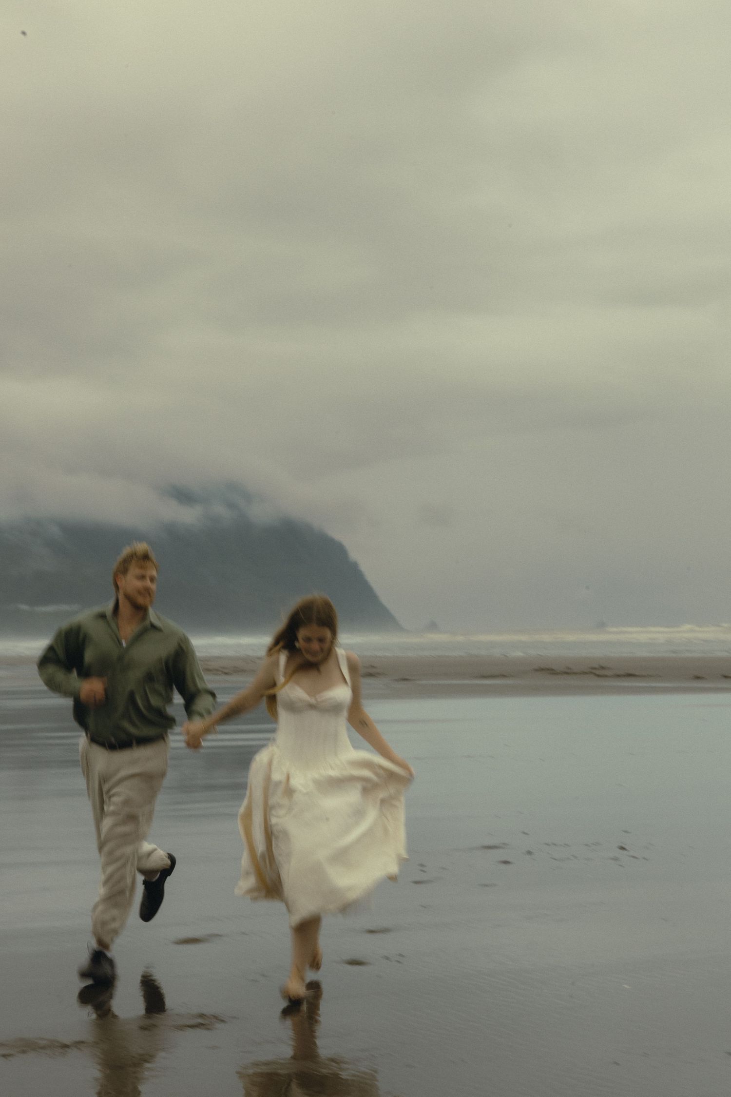 A couple in wedding attire runs joyfully along a misty beach with mountains in the background.