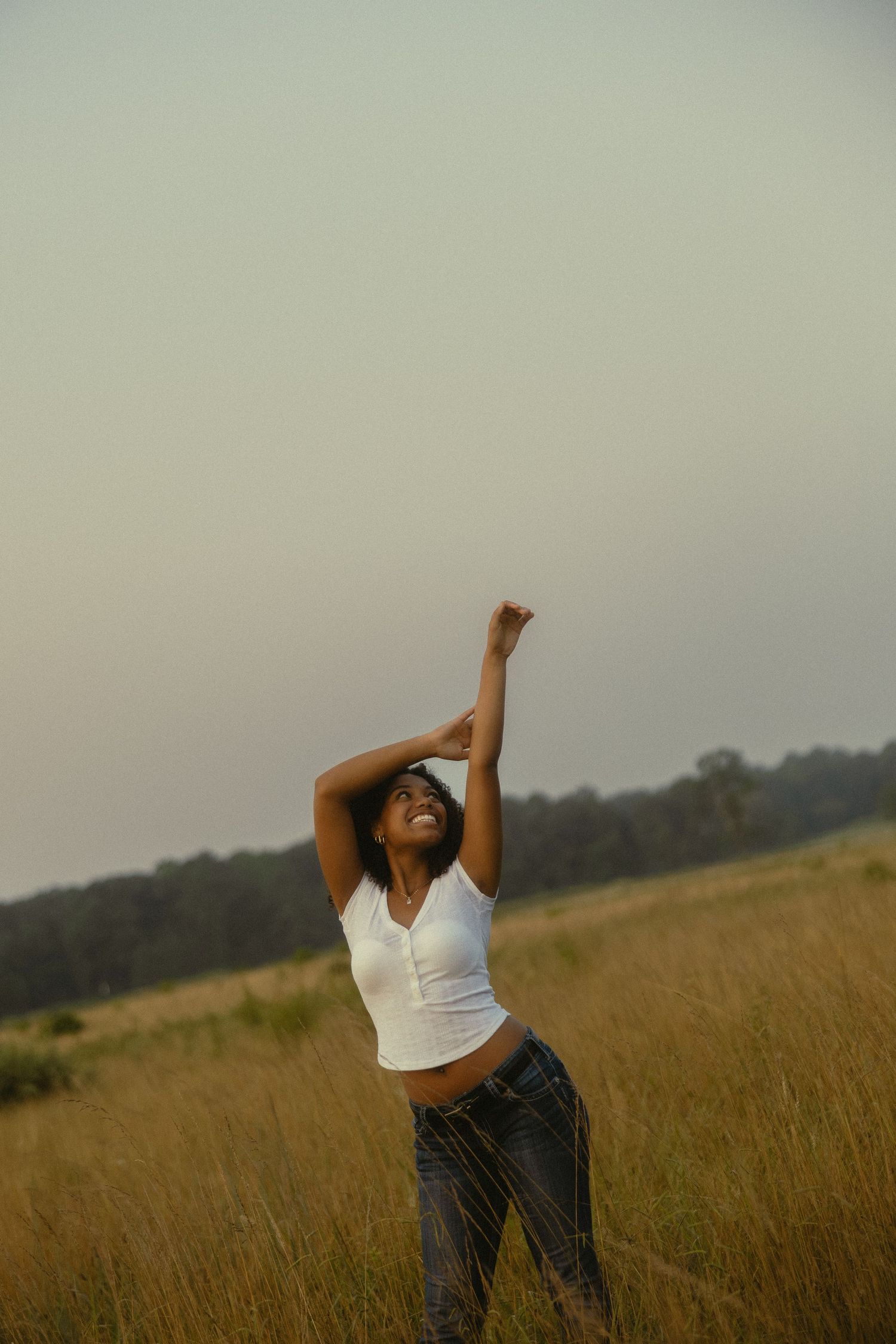 A joyful young Black woman stretches her arms overhead in a golden meadow, wearing a white top and jeans at dusk.