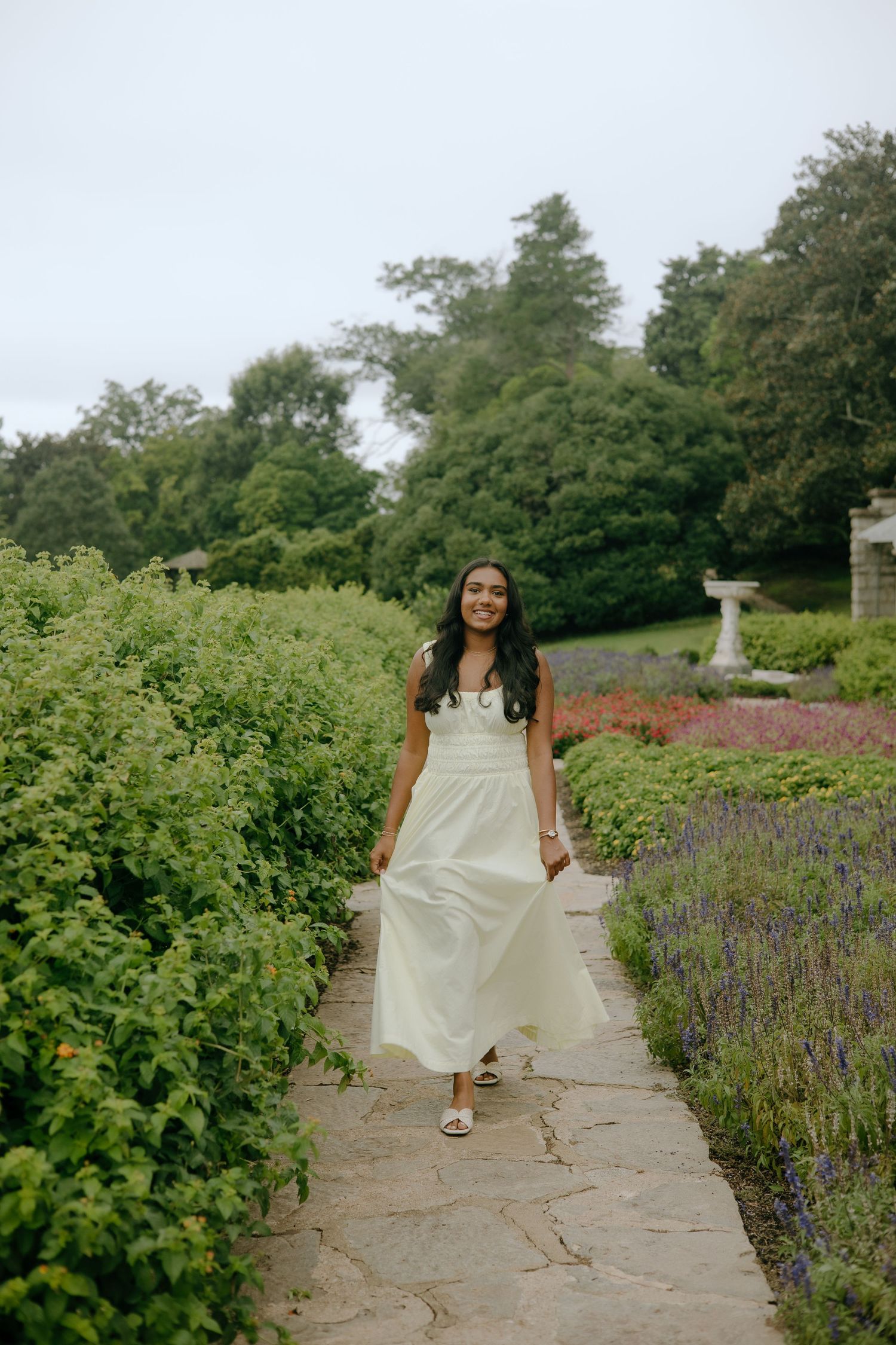 A young woman in a flowing white maxi dress walks along a stone garden path surrounded by lush greenery.