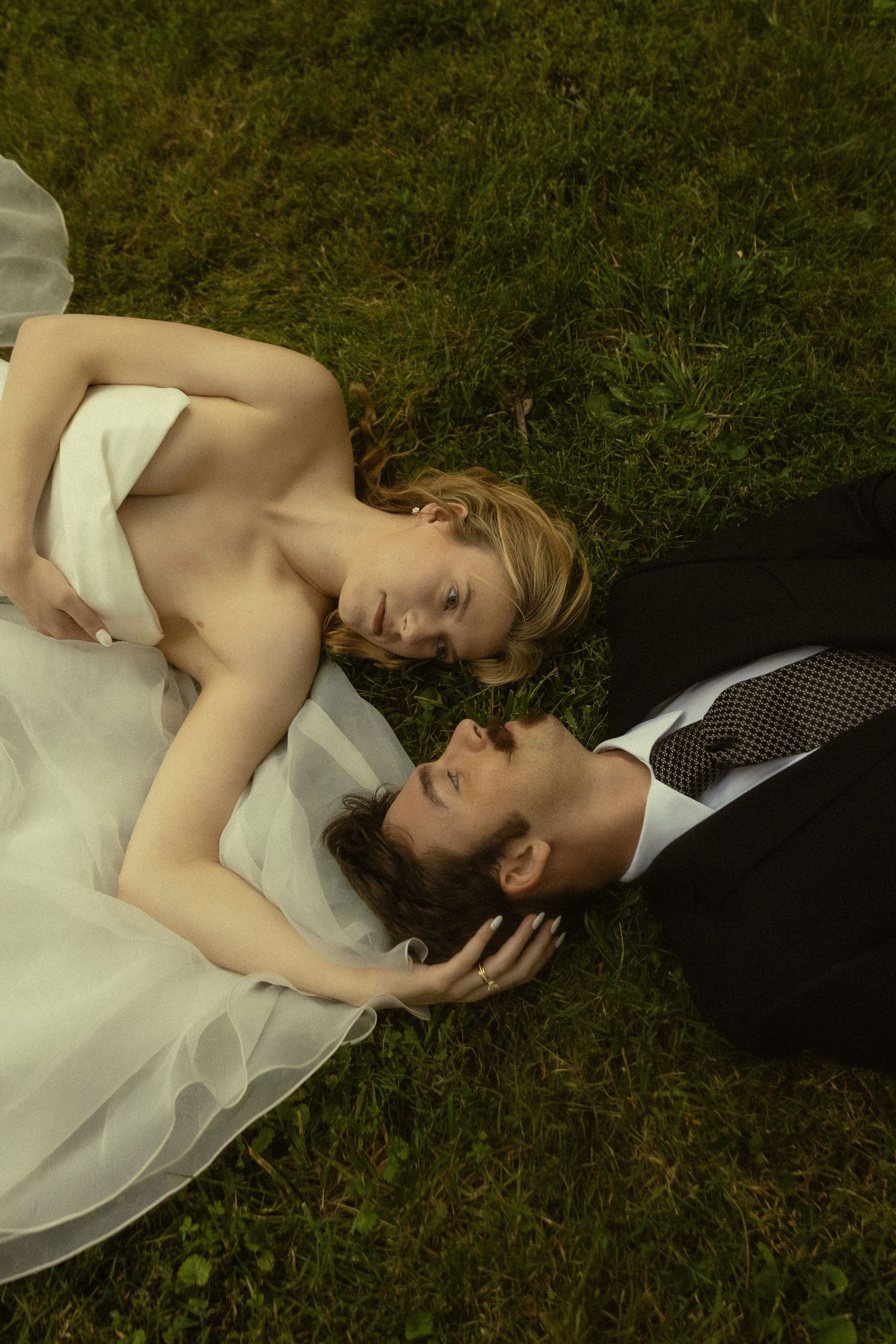 Bride and groom in wedding attire lying on green grass, gazing at each other in a romantic outdoor portrait.