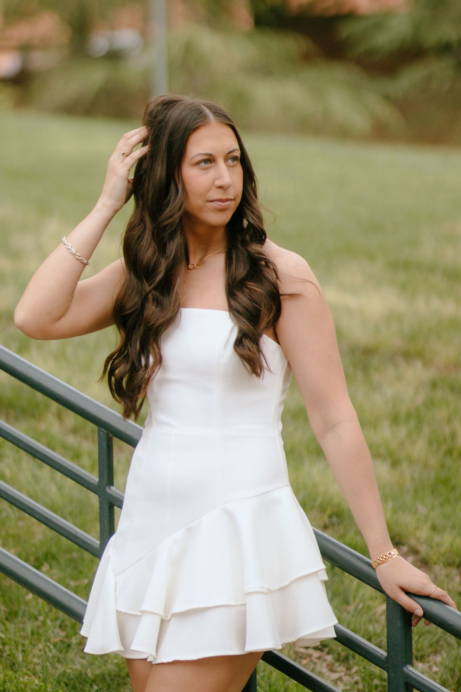 Graduate in white dress poses by metal railing during outdoor photo session.