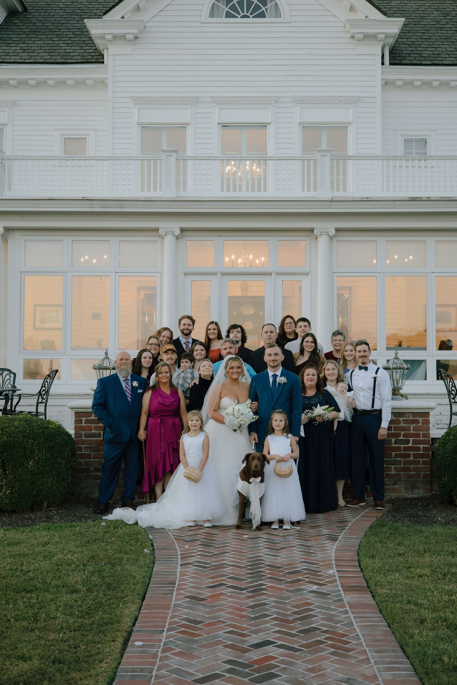 Wedding party and guests pose for a group photo outside a grand white colonial-style venue.