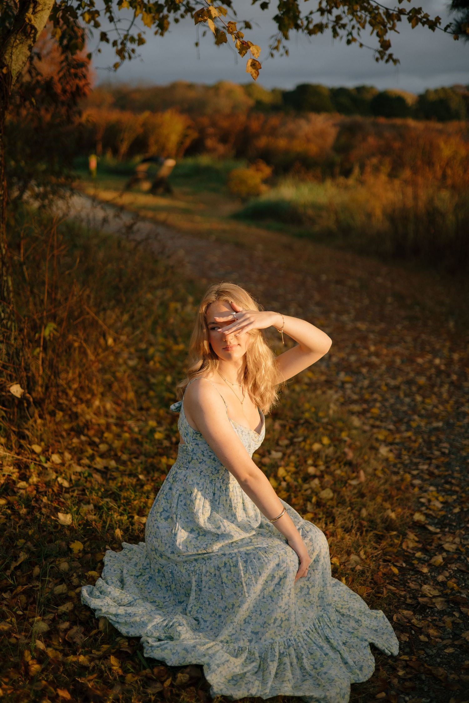 A person in a light blue dress sits gracefully among autumn leaves during golden hour sunset.