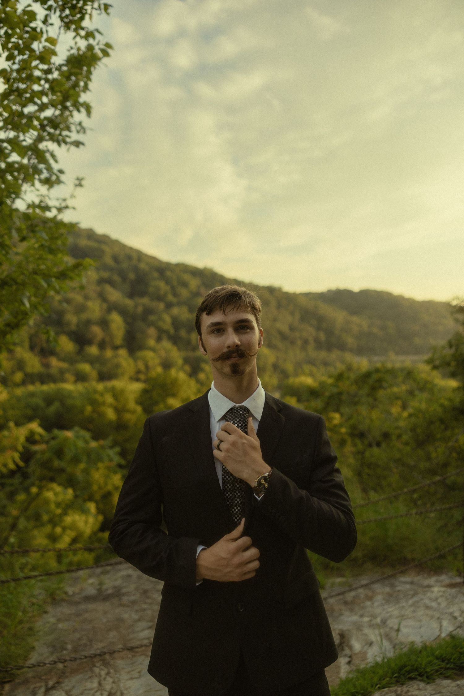Dapper groom with mustache adjusting tie in black suit against scenic green hillside backdrop.