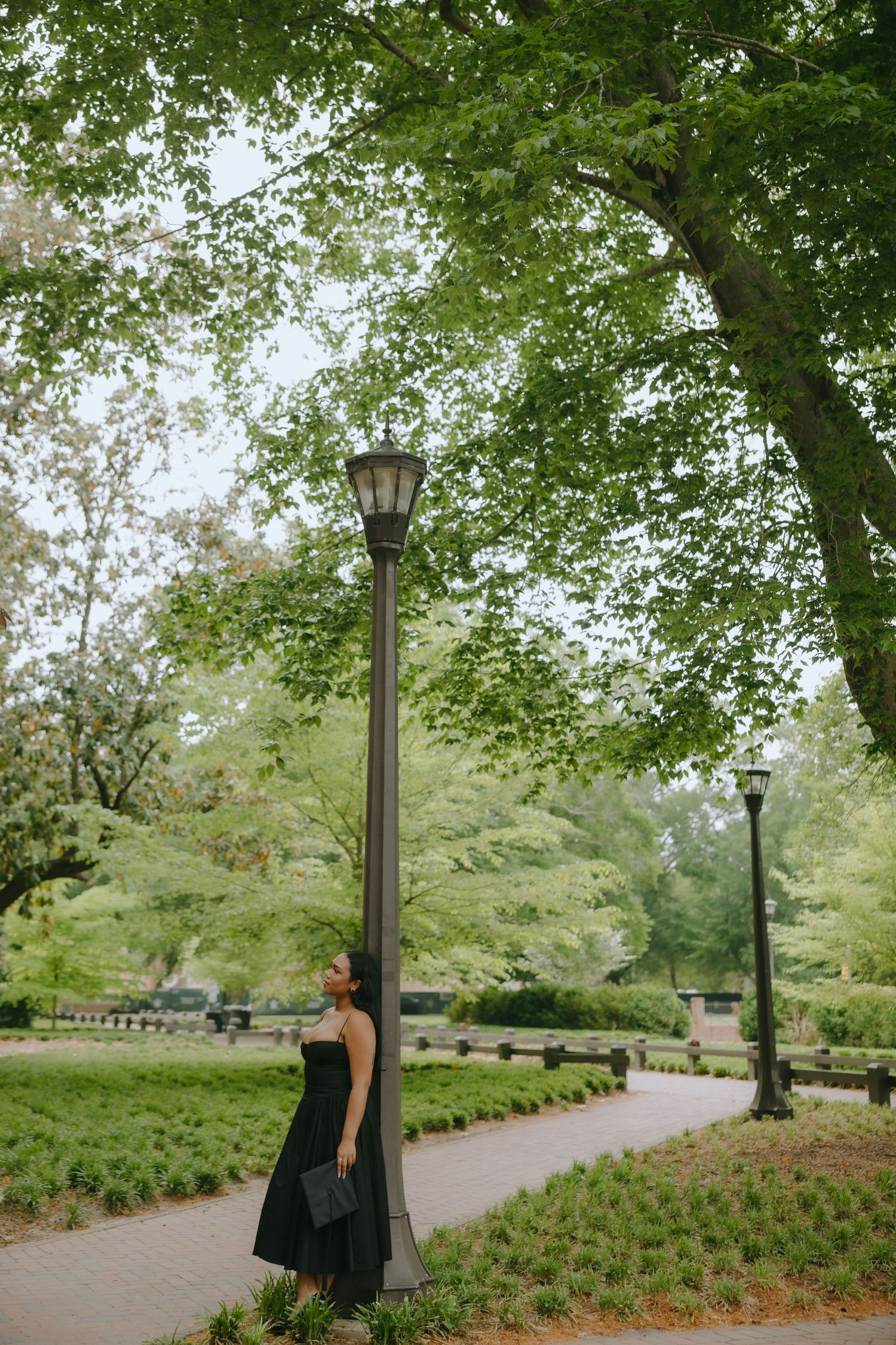 Woman in black dress leaning against a lamp post on a scenic park pathway surrounded by lush green trees.