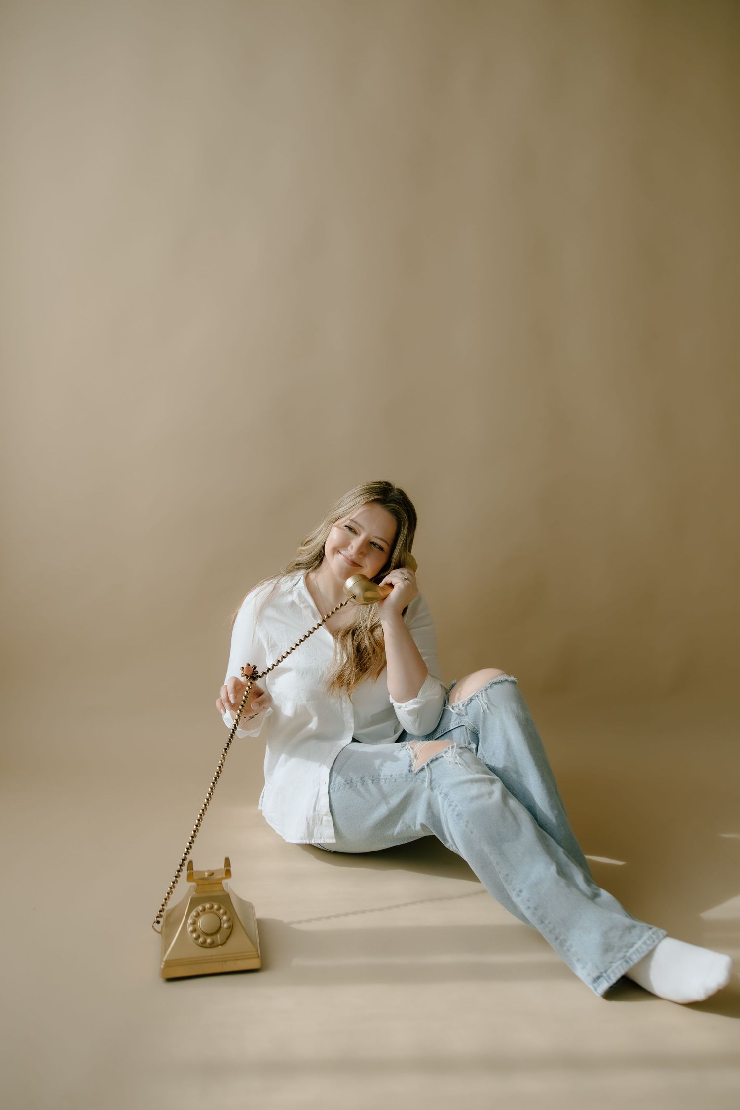 Smiling woman in white shirt and jeans sitting on floor talking on vintage rotary phone against beige background.