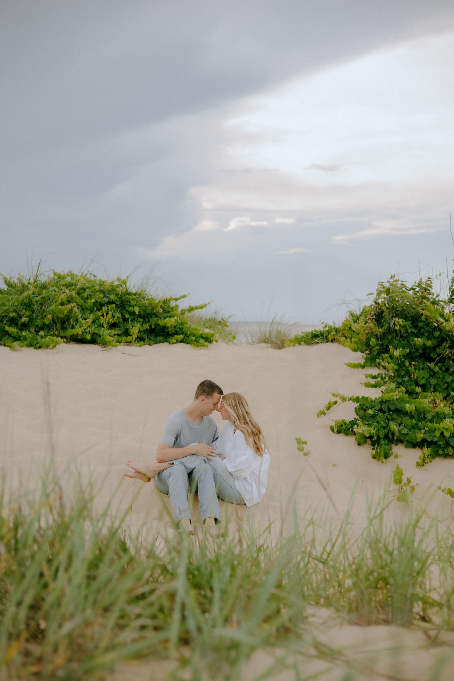 Couple sharing a romantic moment on sandy dunes surrounded by green shrubs under a cloudy sky.
