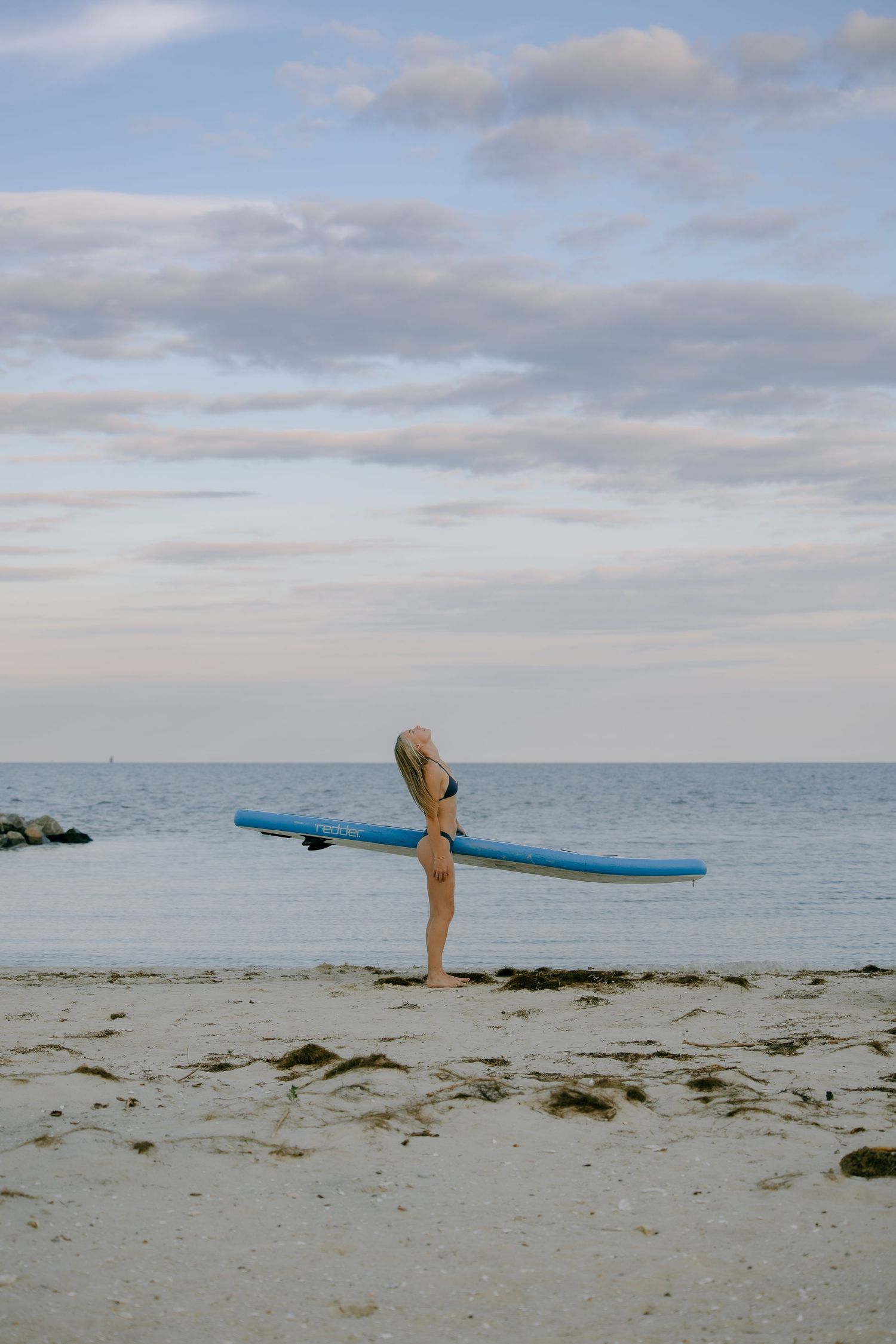 Woman in bikini carrying a blue surfboard on a sandy beach at sunset with calm ocean waters in the background.