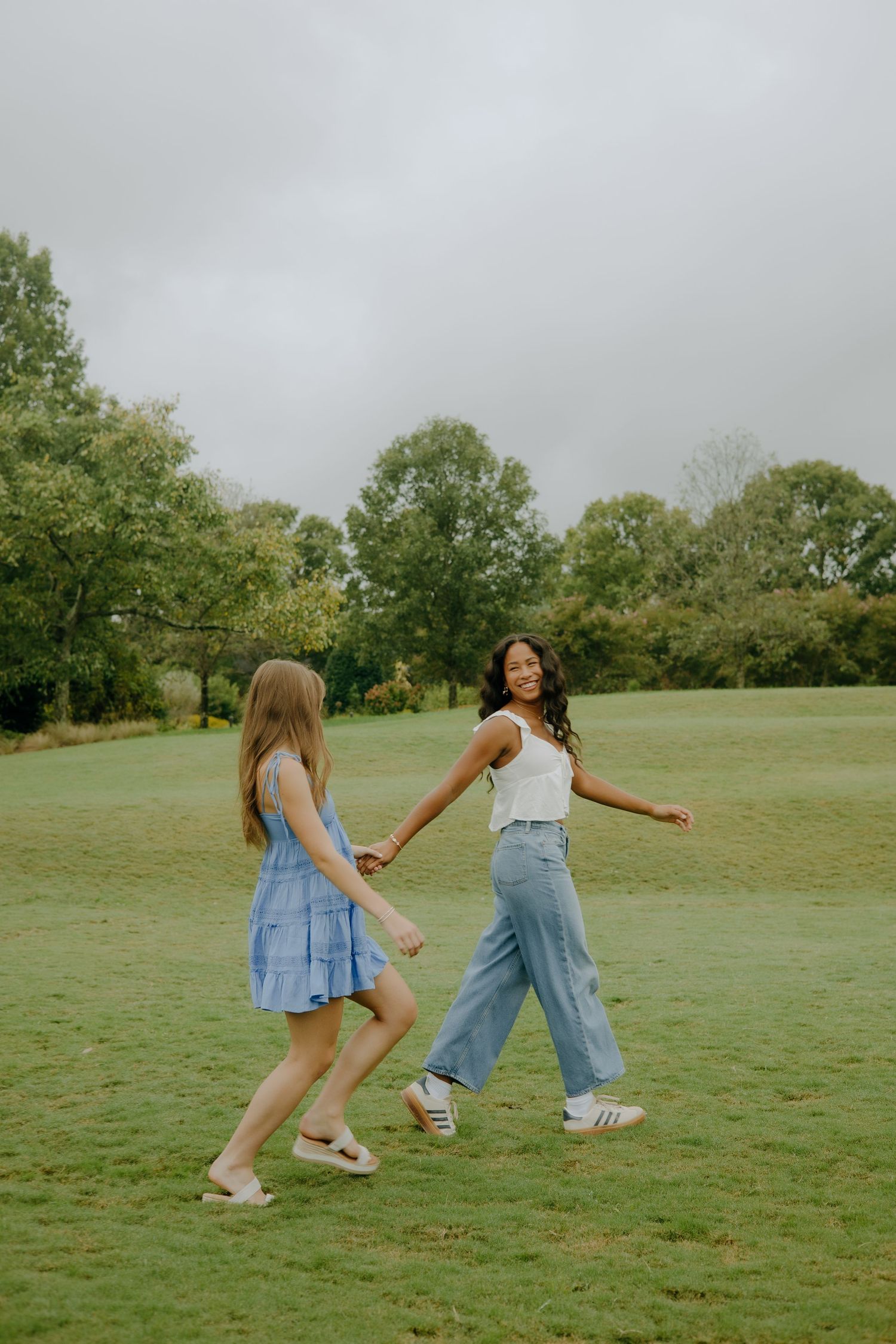 Two young women holding hands and walking together on a green grass field with trees in the background.