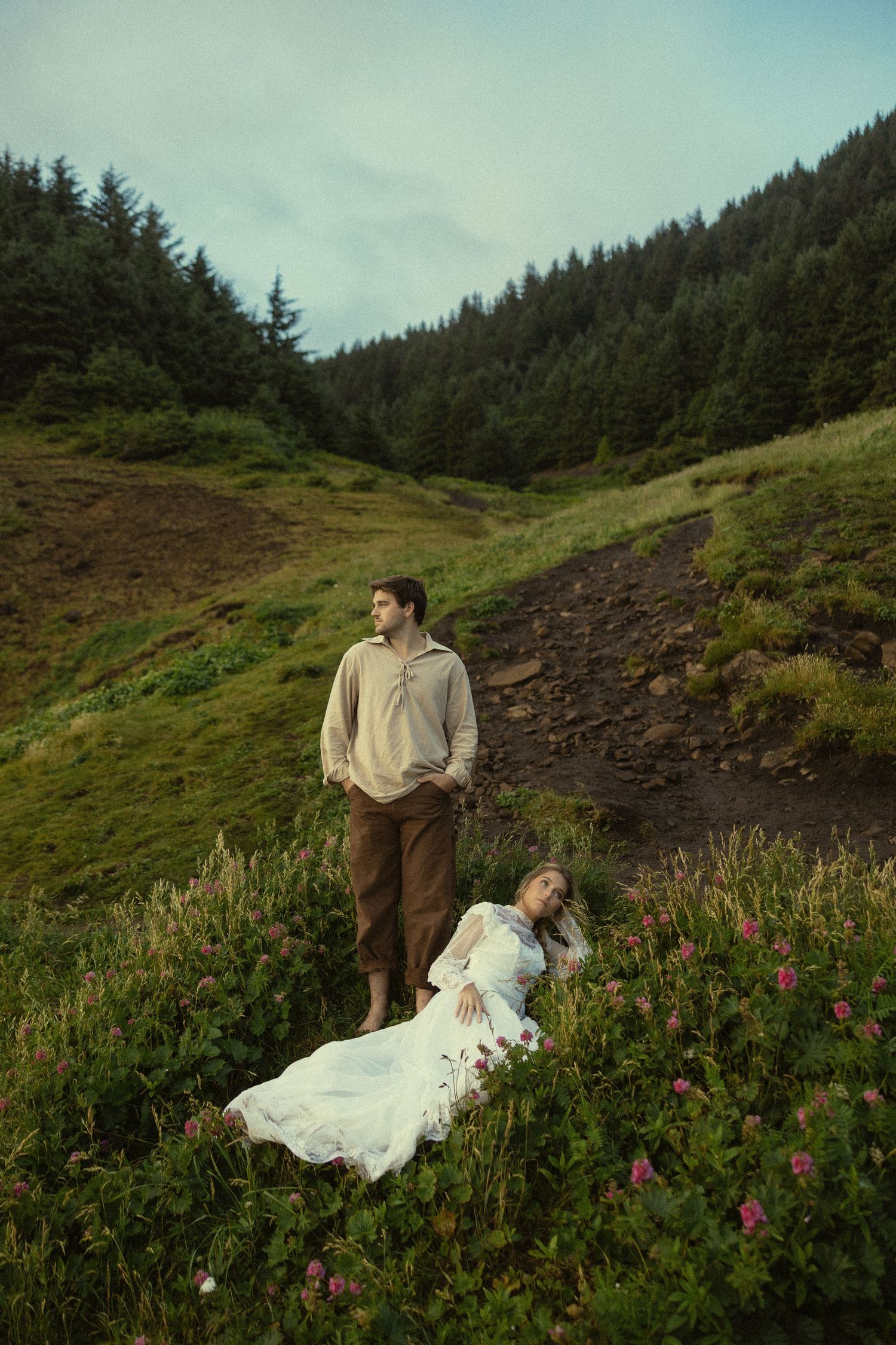 Man in beige hoodie stands on hillside trail as woman in white dress lies among pink wildflowers.