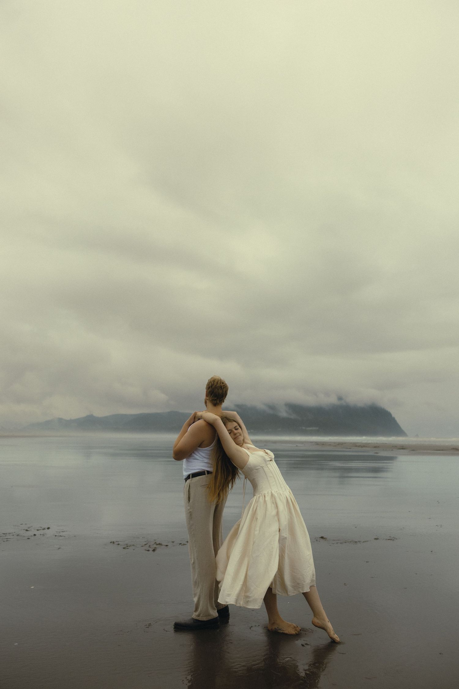 A couple embraces on a misty beach, the woman in a white dress leaning into her partner with mountains in the background.