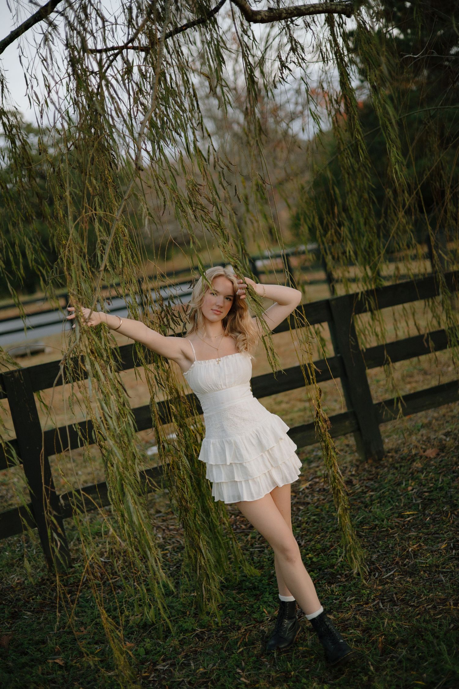A figure in a white dress dances gracefully among willow branches and wooden fences in a natural outdoor setting.