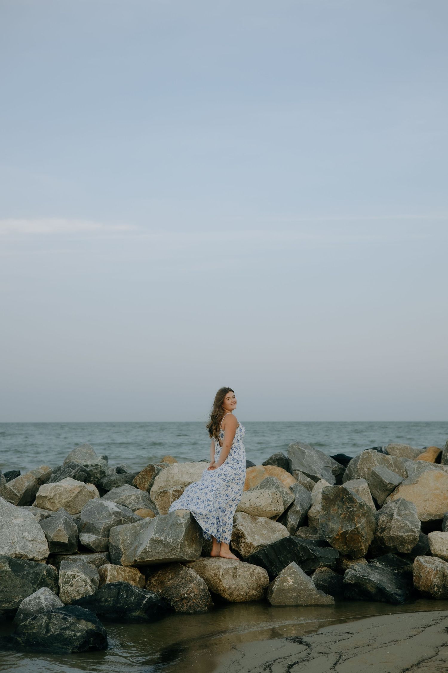 A woman in a white floral dress sits peacefully on coastal rocks at sunset by the ocean.