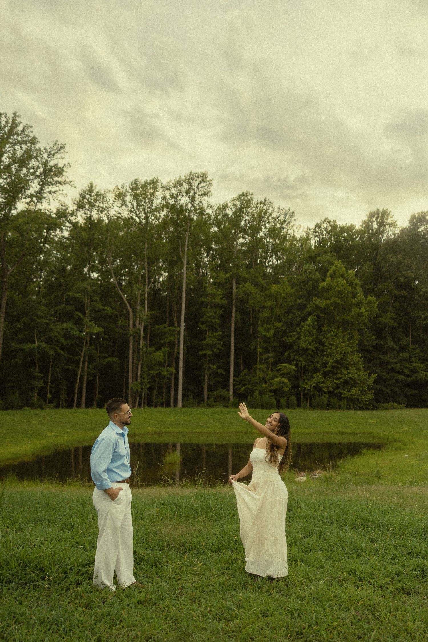 Couple in white dress and blue shirt dancing in lush green field near pond with tall trees backdrop.