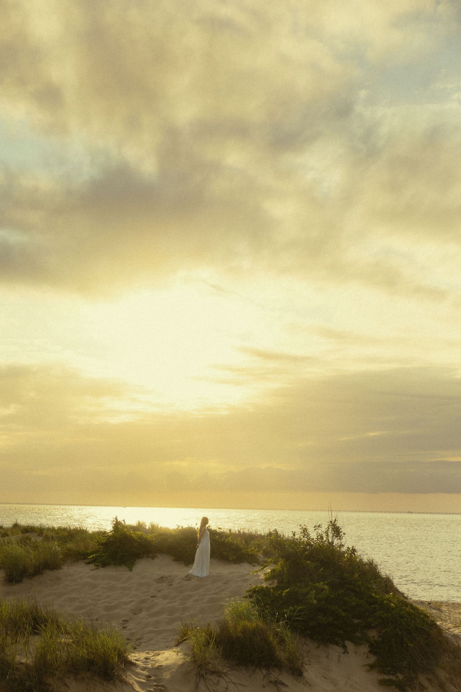 A woman in a white dress stands on sandy dunes overlooking a golden sunset over the ocean.