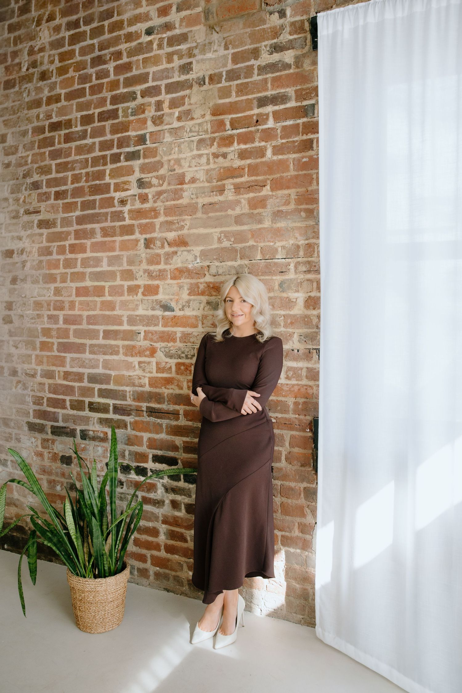 Woman in brown long sleeve dress poses against exposed brick wall with potted plant and white curtain.