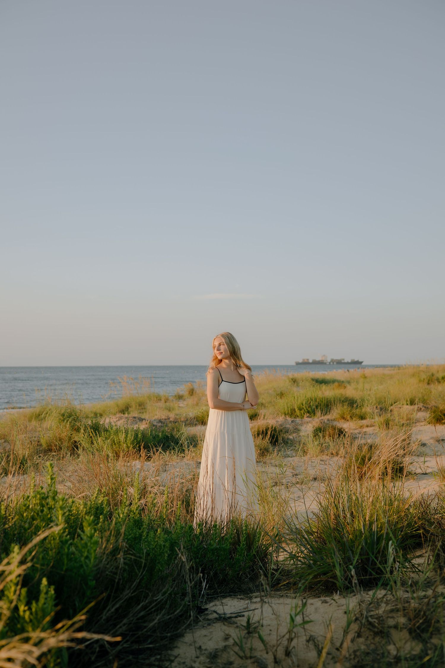 Young blonde woman in white skirt stands on sandy beach dunes with ocean and ship in background at sunset.
