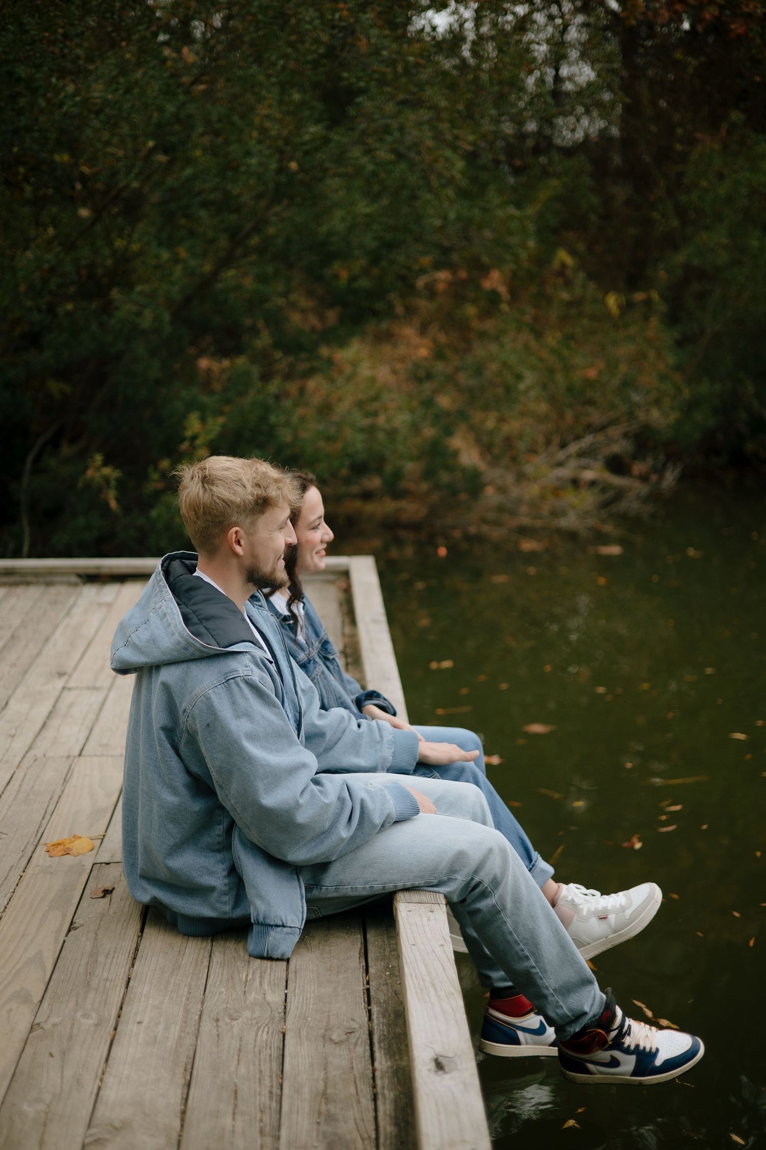 Couple sitting on a wooden dock by a calm lake surrounded by autumn foliage.