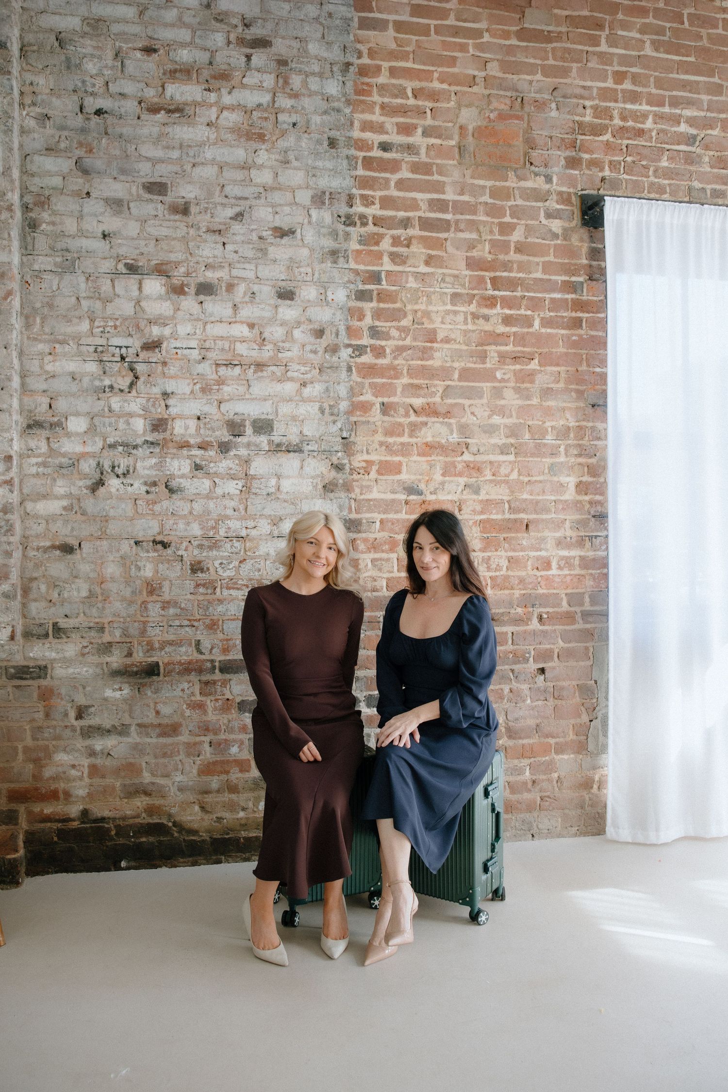 Two elegant women in stylish dresses pose together against a rustic brick wall with a green suitcase.